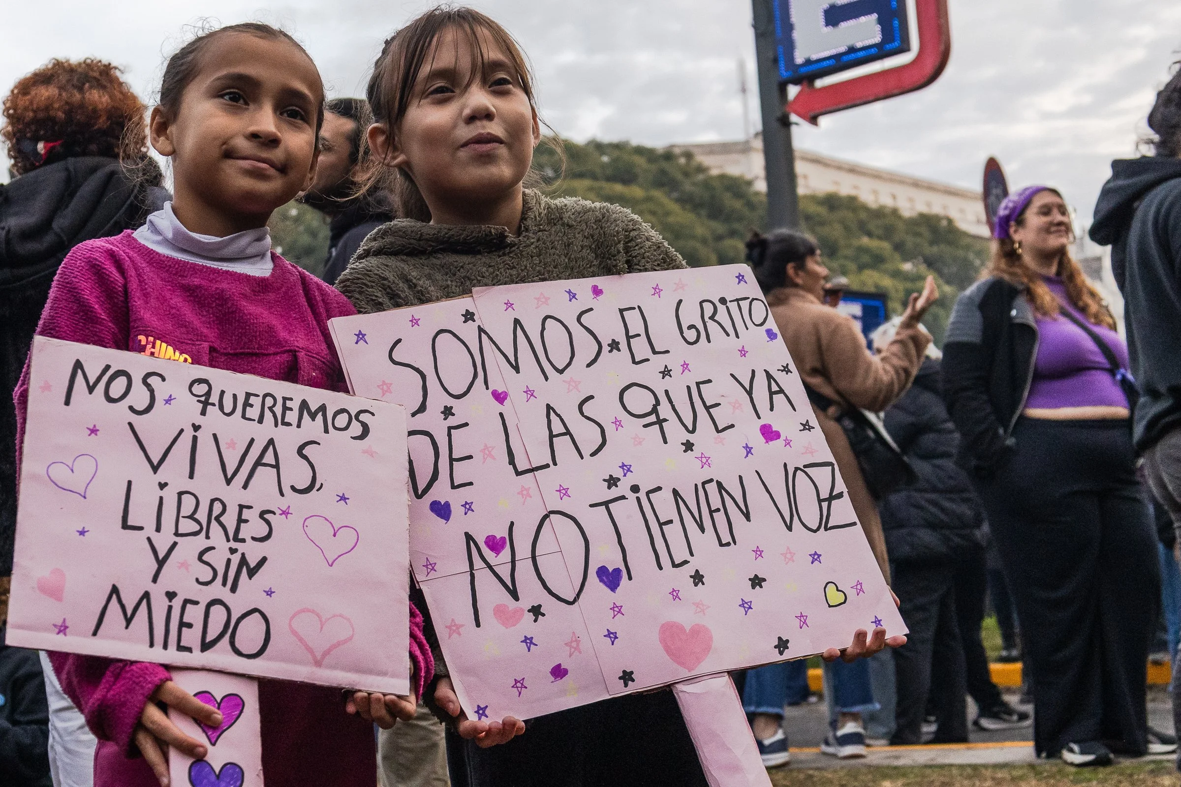 Dos niñas en protesta sosteniendo carteles con mensajes que dicen "nos queremos vivas, libres y sin miedo" y "somos el gritos de las que ya no están", rodeadas de otras personas en un espacio al aire libre con cielo nublado.