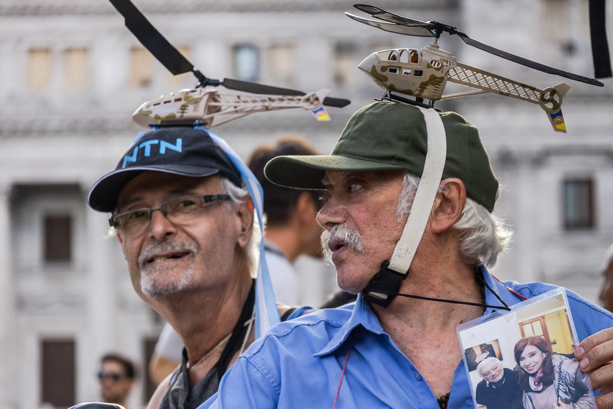 Hombres mayores con cascos de protección y una helicóptero de juguete en la cabeza, uno de ellos sostiene una foto
