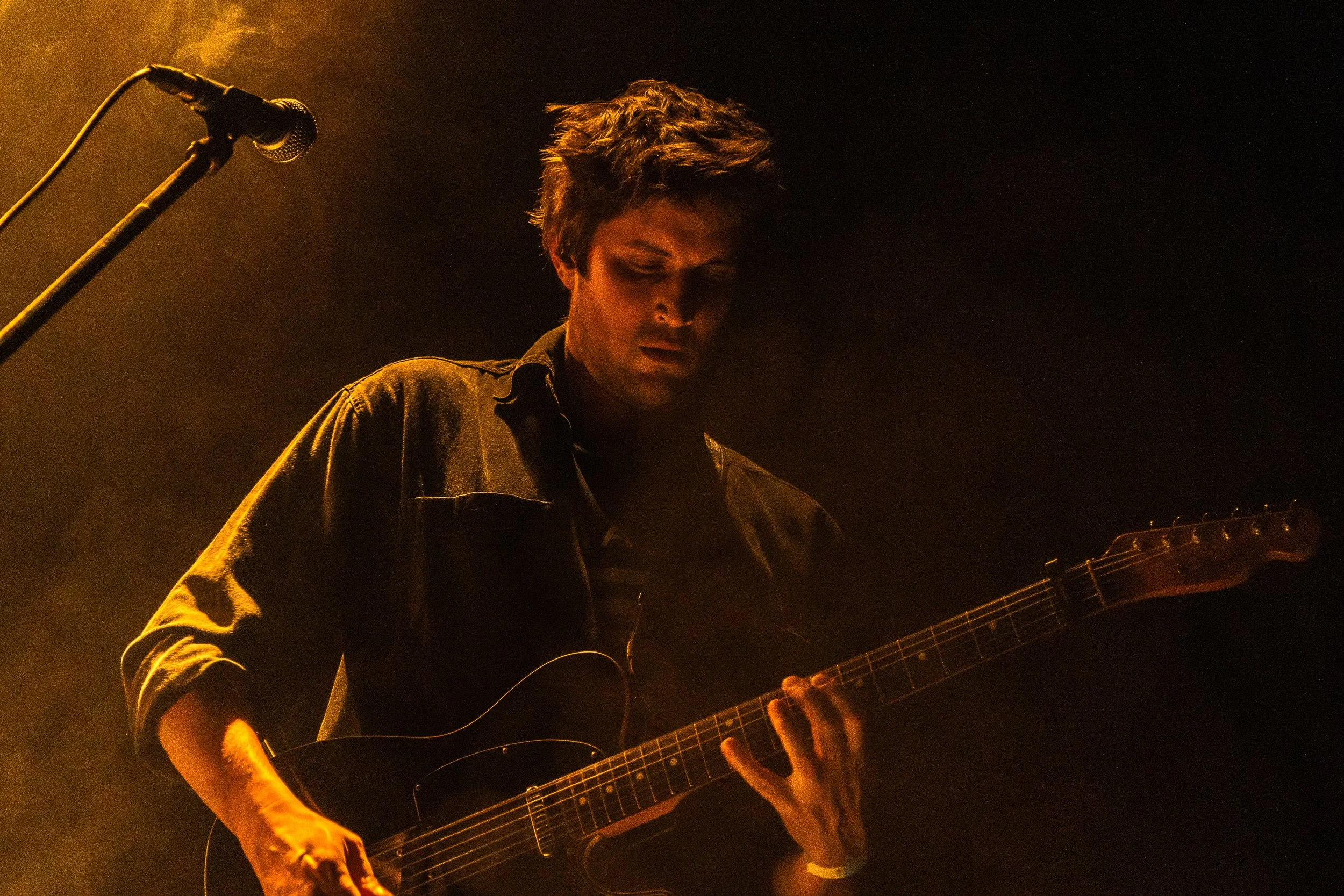 Joven tocando guitarra acústica en un escenario con iluminación cálida y fondo oscuro.