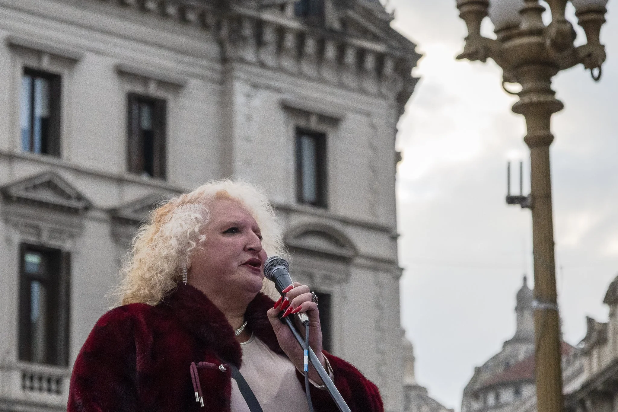 Mujer de cabello rubio rizado hablando por micrófono en una plaza con edificios antiguos y un poste de luz vintage en el fondo.
