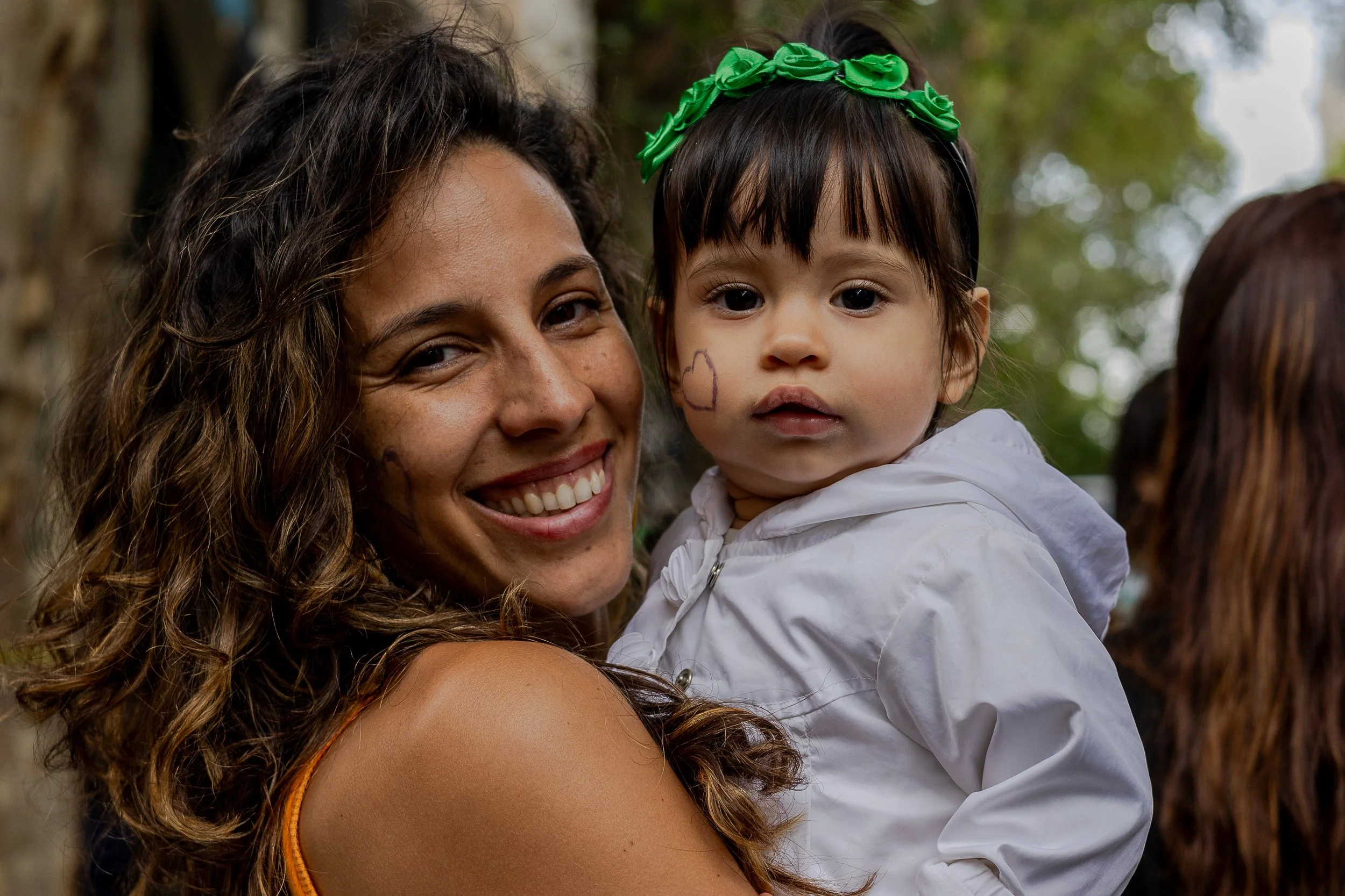 Mujer sonriente sosteniendo a una niña pequeña con risa y decoraciones en la cara en un ambiente al aire libre