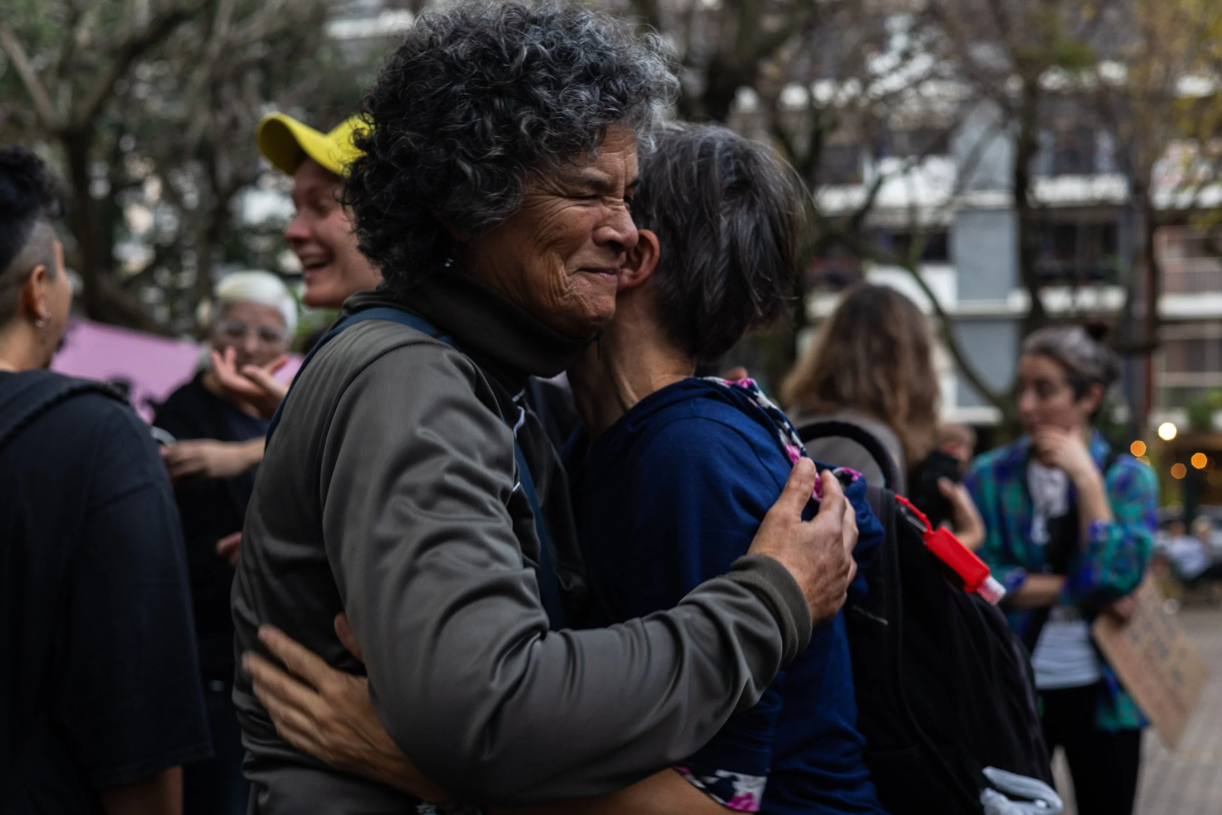 Dos personas se abrazan en una reunión al aire libre con varias otras personas en el fondo, en un ambiente que parece ser un evento social o protesta.