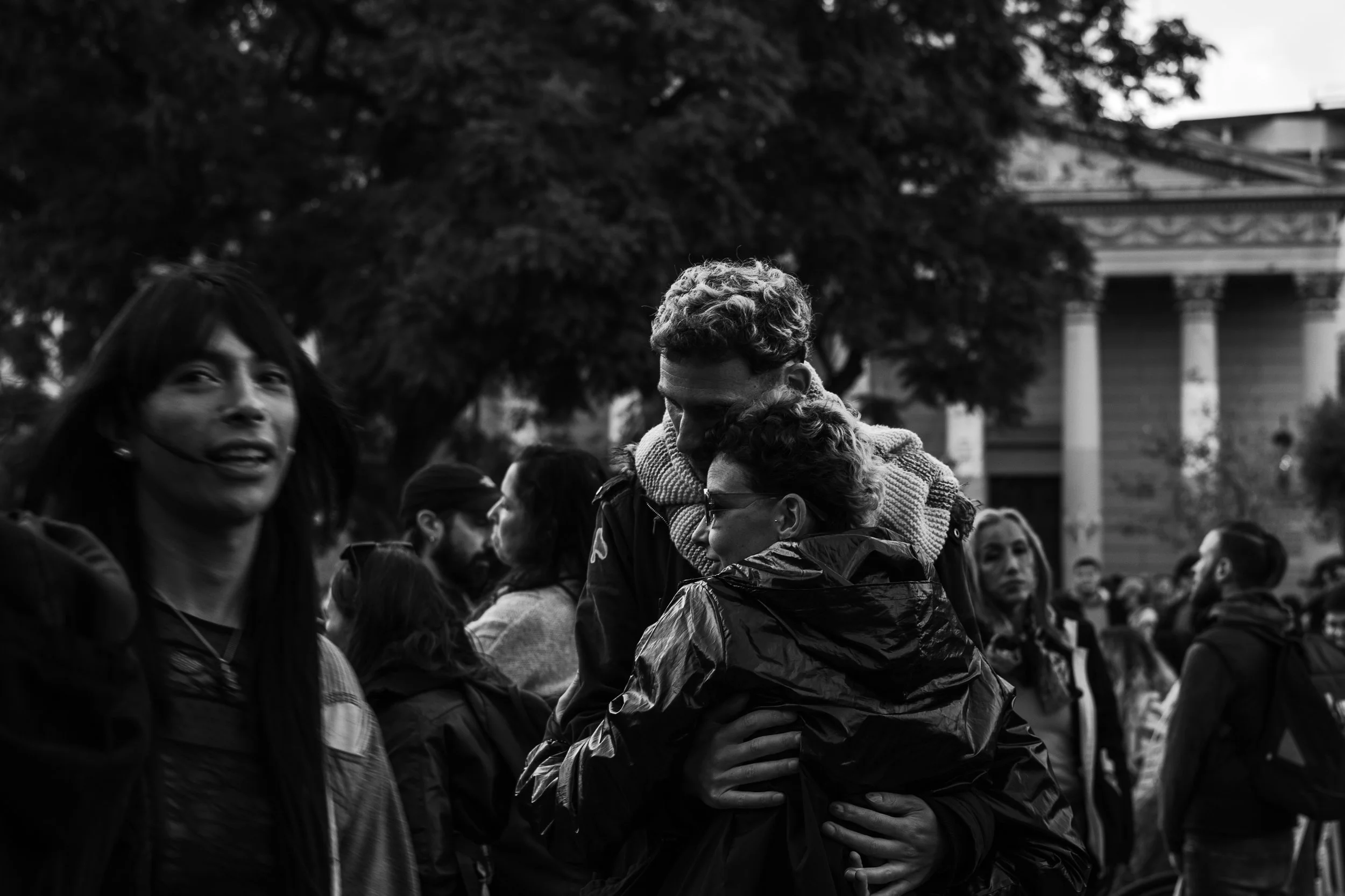 Grupo de personas en un espacio público, algunas se abrazan y otras conversan, en un entorno con árboles y una estructura clásica en el fondo, en tonos negros y blancos.
