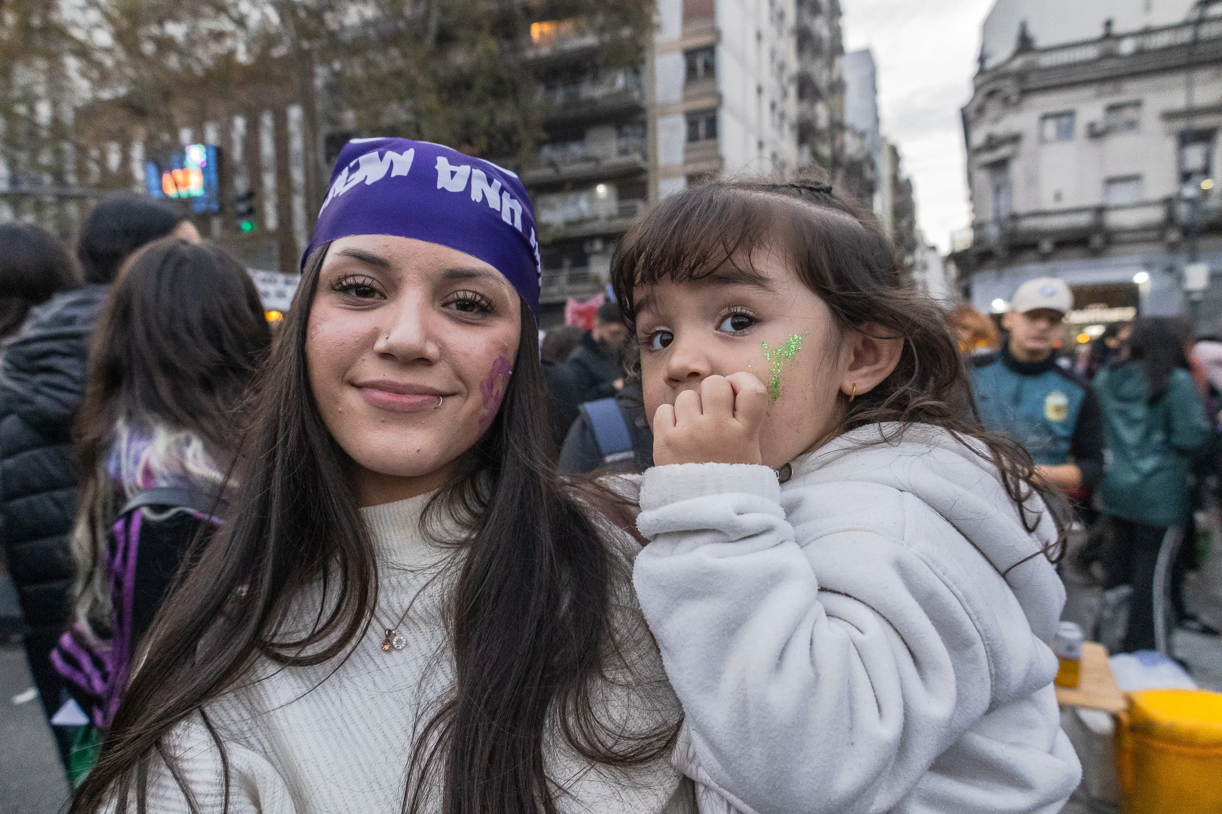 Mujer y niña con pintura de cara en la calle durante una manifestación o evento en ciudad, con edificios de fondo y muchas personas.
