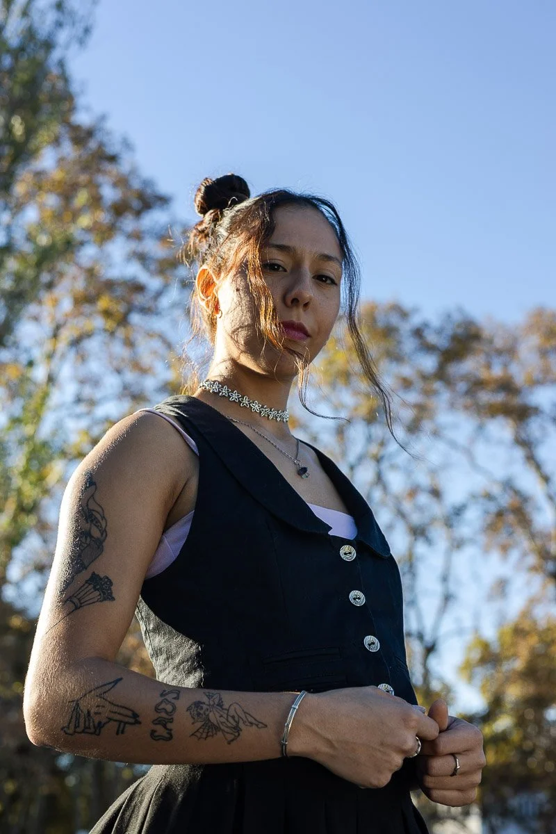 Juvenil mujer con tatuajes, peinado en moño, gafas negras, vestido negro sin mangas, en un parque con árboles de fondo y cielo despejado.
