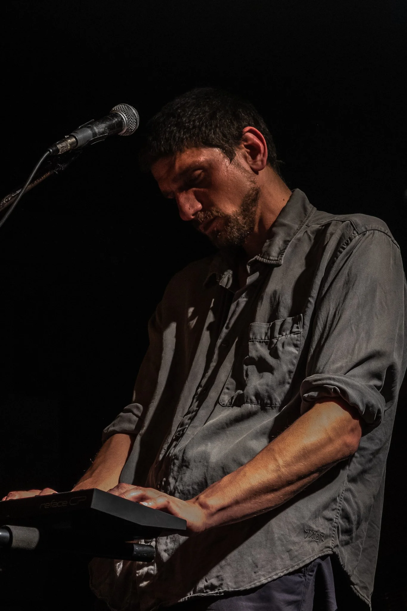Hombre tocando un teclado en un escenario oscuro, con micrófono frente a él.