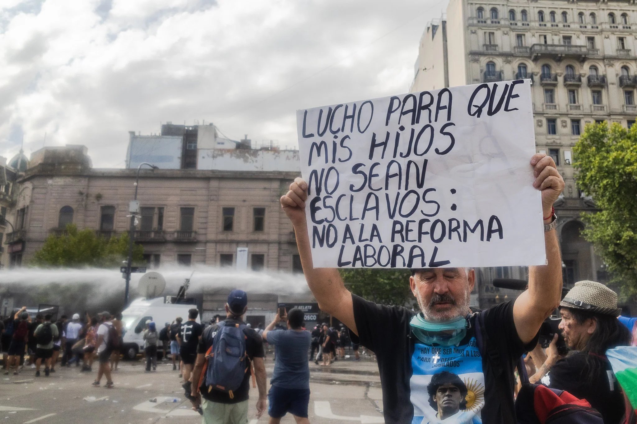 Persona en una manifestación sostiene cartel con texto que denuncia la explotación de hijos, con edificios y un grupo de personas en el fondo.
