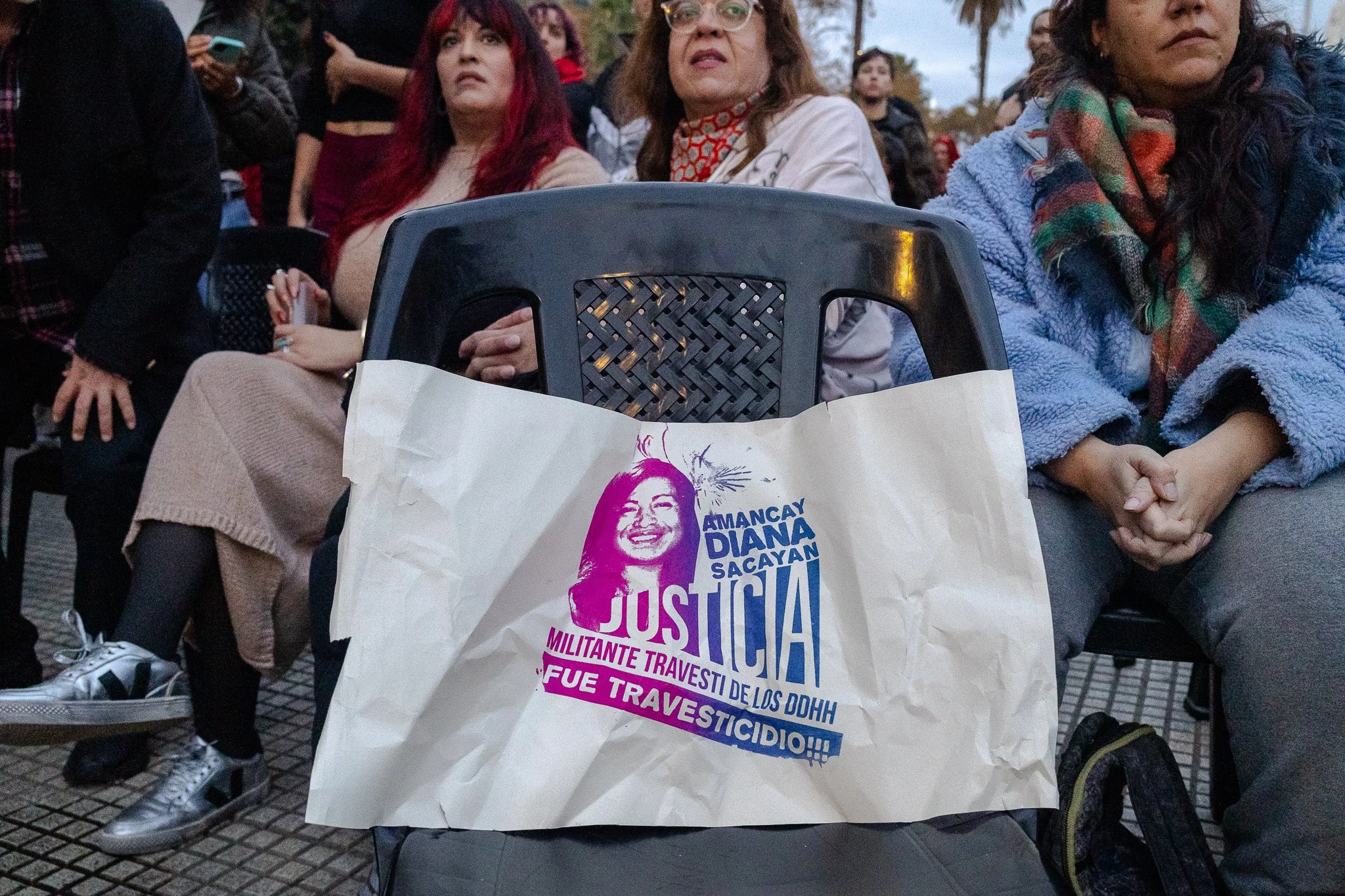 Personas sentadas en un evento, una silla en primer plano con un cartel que dice "Justicia" con la cara de la activista trans Diana Sacayán.