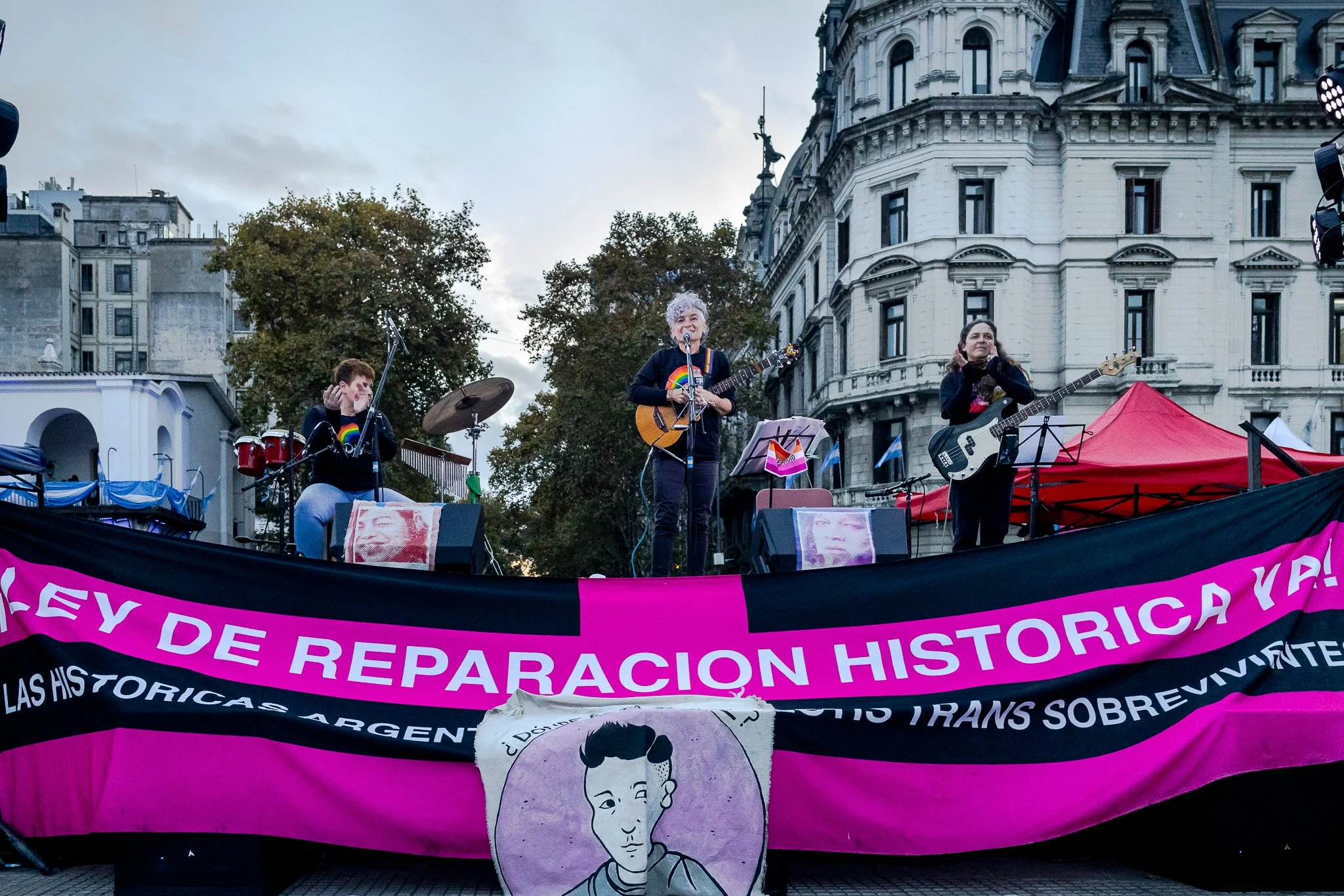 Una manifestación en una plaza con personas tocando instrumentos y un cartel grande en primer plano que dice 'Ley de Reparación Histórica'.
