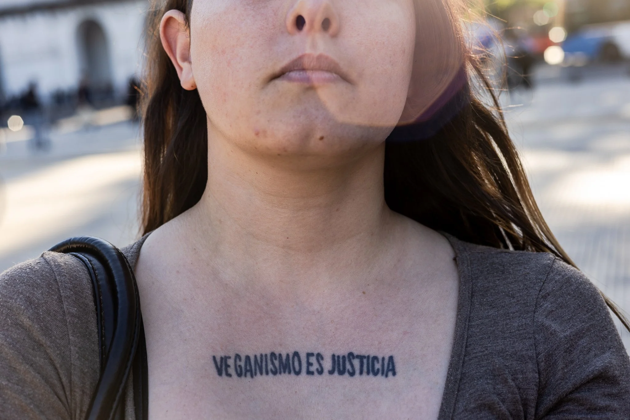 Mujer con un tatuaje en el pecho que dice 'Veganismo es justicia' en letras negras, en un entorno exterior con personas y autos en el fondo.
