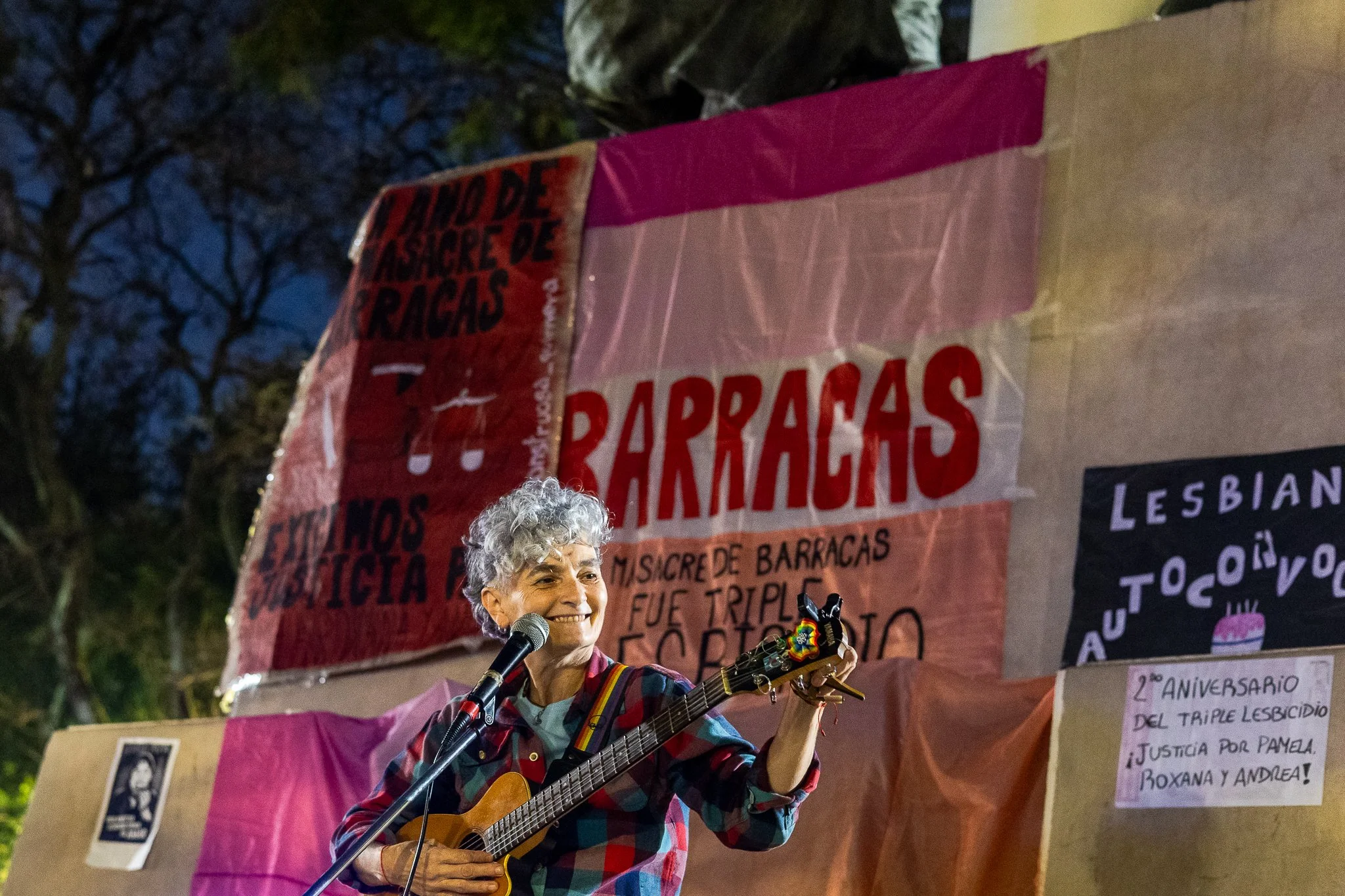 Una mujer mayor tocando la guitarra con una sonrisa, en un escenario de protesta con pancartas de fondo. Es de noche y hay árboles y pancartas con mensajes relacionados con la lucha y justicia.