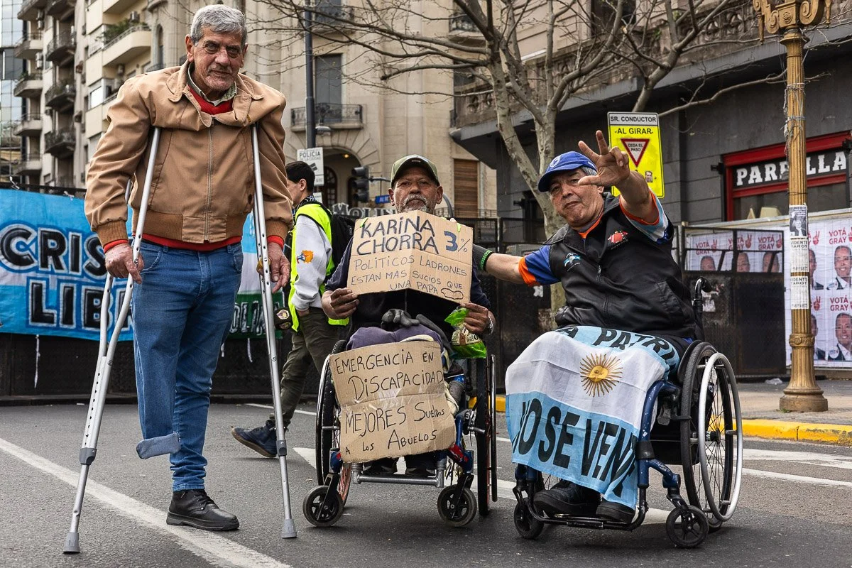 Dos hombres en sillas de ruedas en una protesta en la calle, uno con una bandera argentina y el otro con carteles. La escena muestra una manifestación en una ciudad.