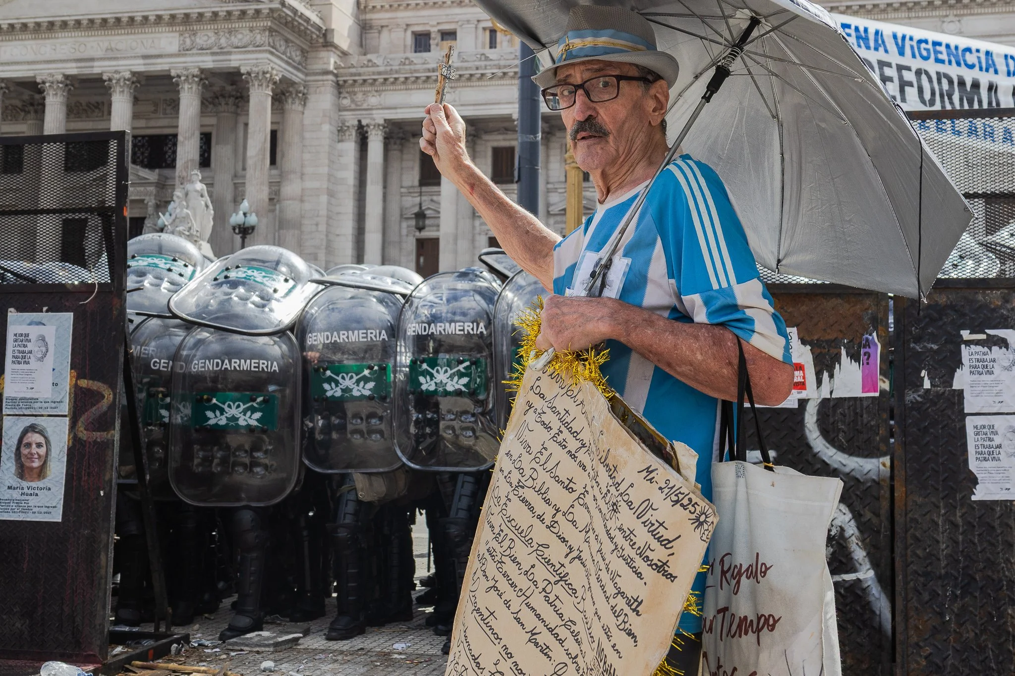Un hombre mayor con sombrero, gafas y camiseta azul y blanca sostiene una cruz en una mano y un cartel pintado a mano en la otra, en un entorno urbano con edificio antiguo y vallas. Lleva un paraguas y está rodeado de envases de escudo de la policía 