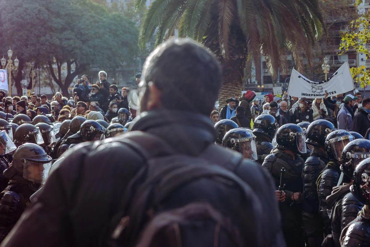 Persona con mochila observando una protesta con muchas personas y policías equipados con cascos en un parque