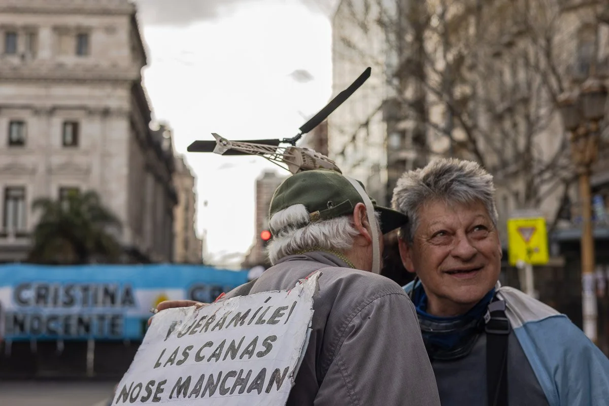 Dos hombres viejos hablando en una calle en el día, uno lleva casco con rotor de helicóptero y una señal en la espalda que dice 'Fueramile! Las canas nos manchan'.