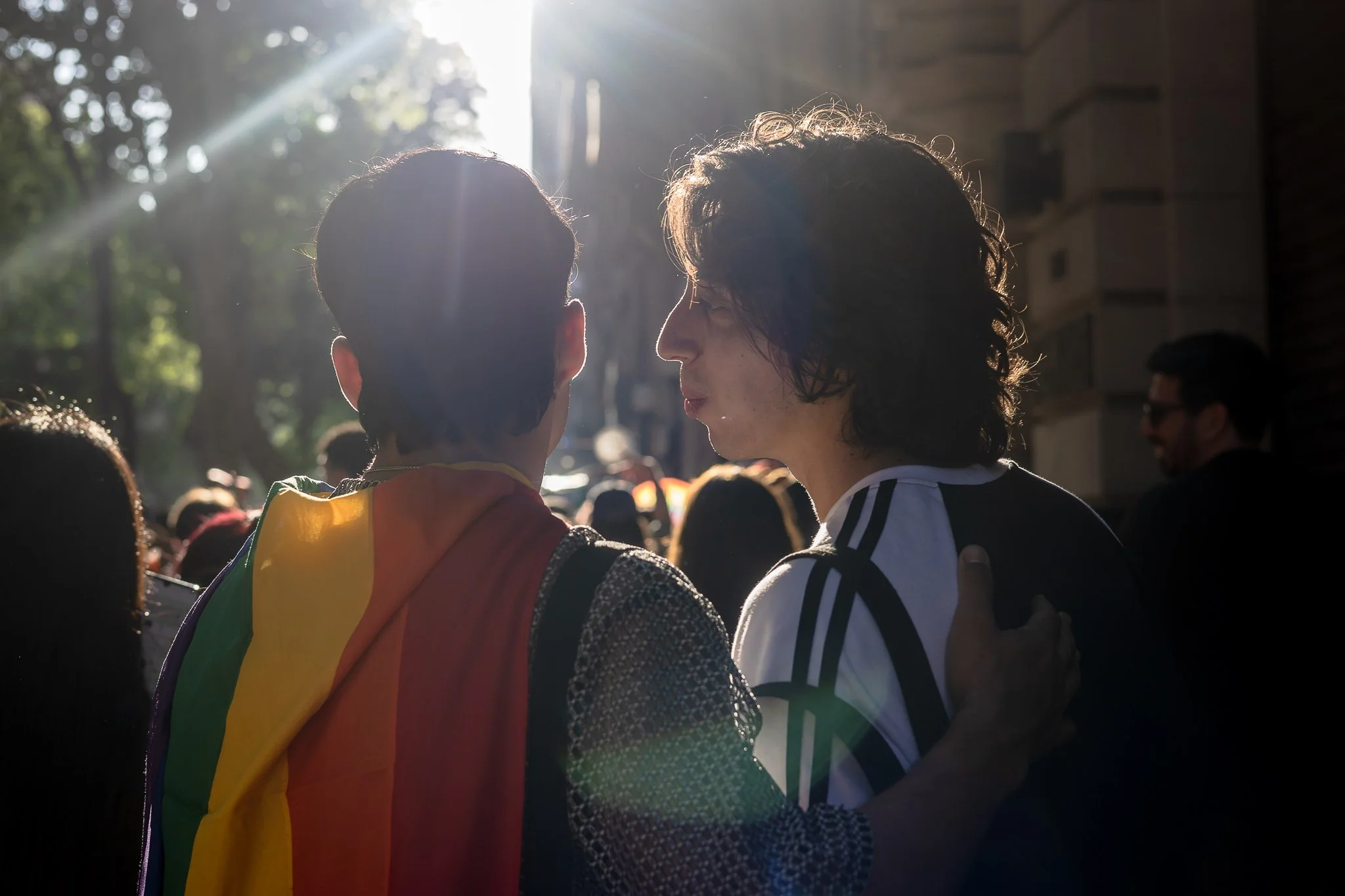 Dos personas conversando en una calle al atardecer, una con una bandera arcoíris y la otra con una camiseta con diseño de círculos