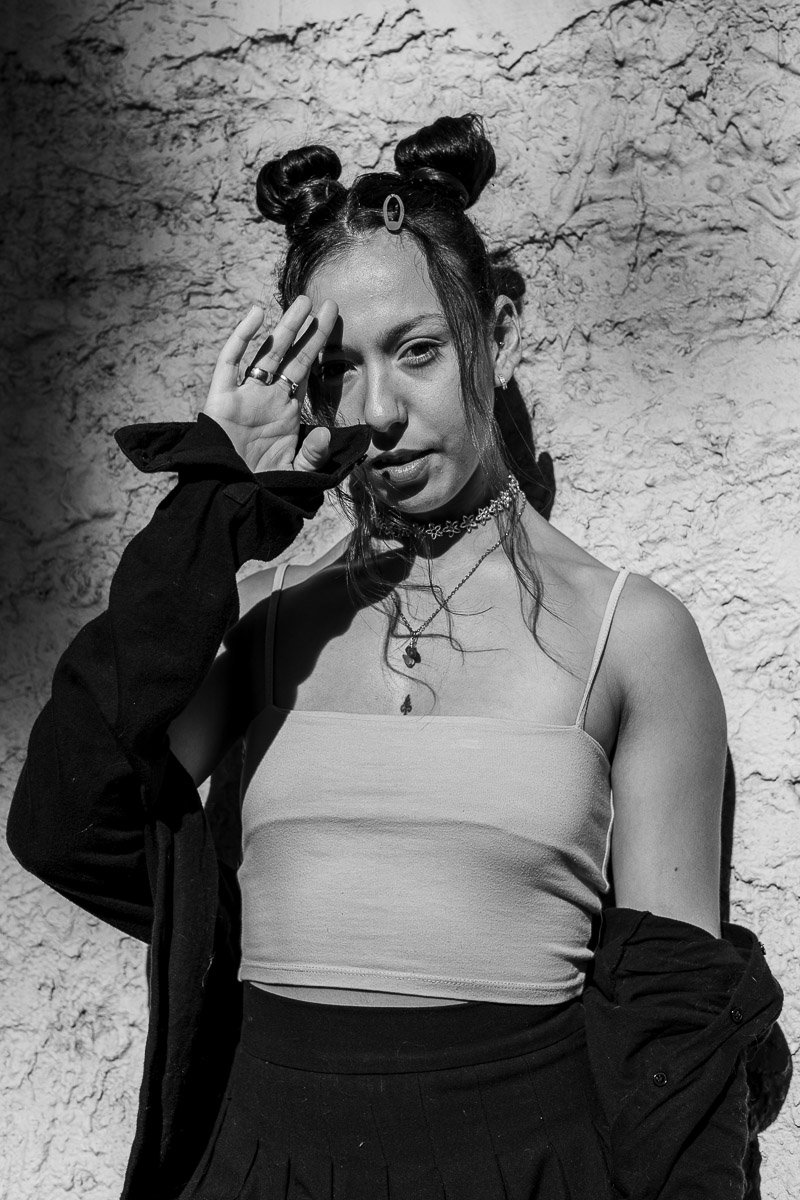 Mujer joven con peinado en dos moños, posando frente a una pared de piedra, con prendas modernas y accesorios, en una fotografía en blanco y negro.