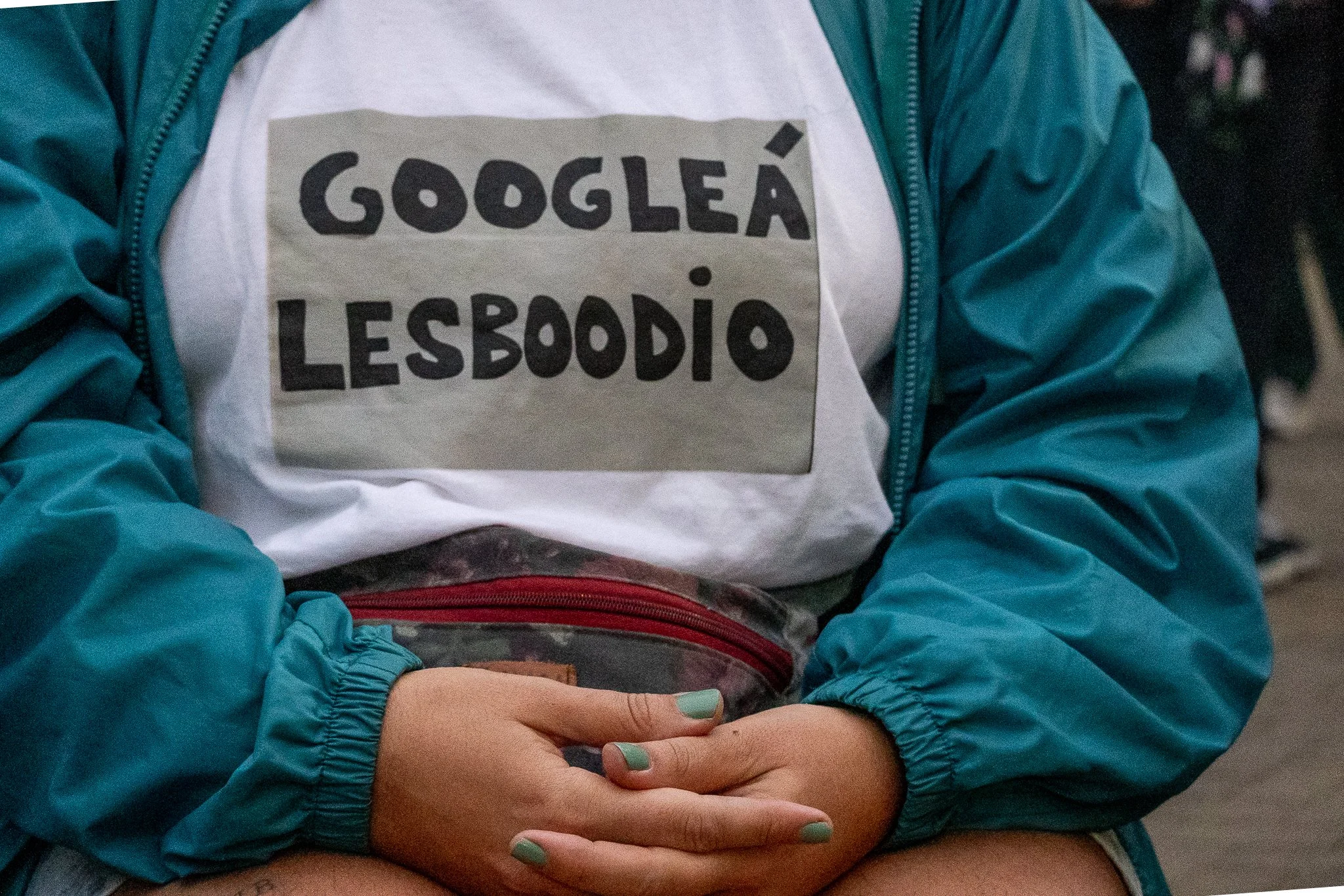Persona con chaqueta azul sosteniendo sus manos en su regazo, usando una camiseta blanca con letras negras que dicen 'Googleá lesboodio'.