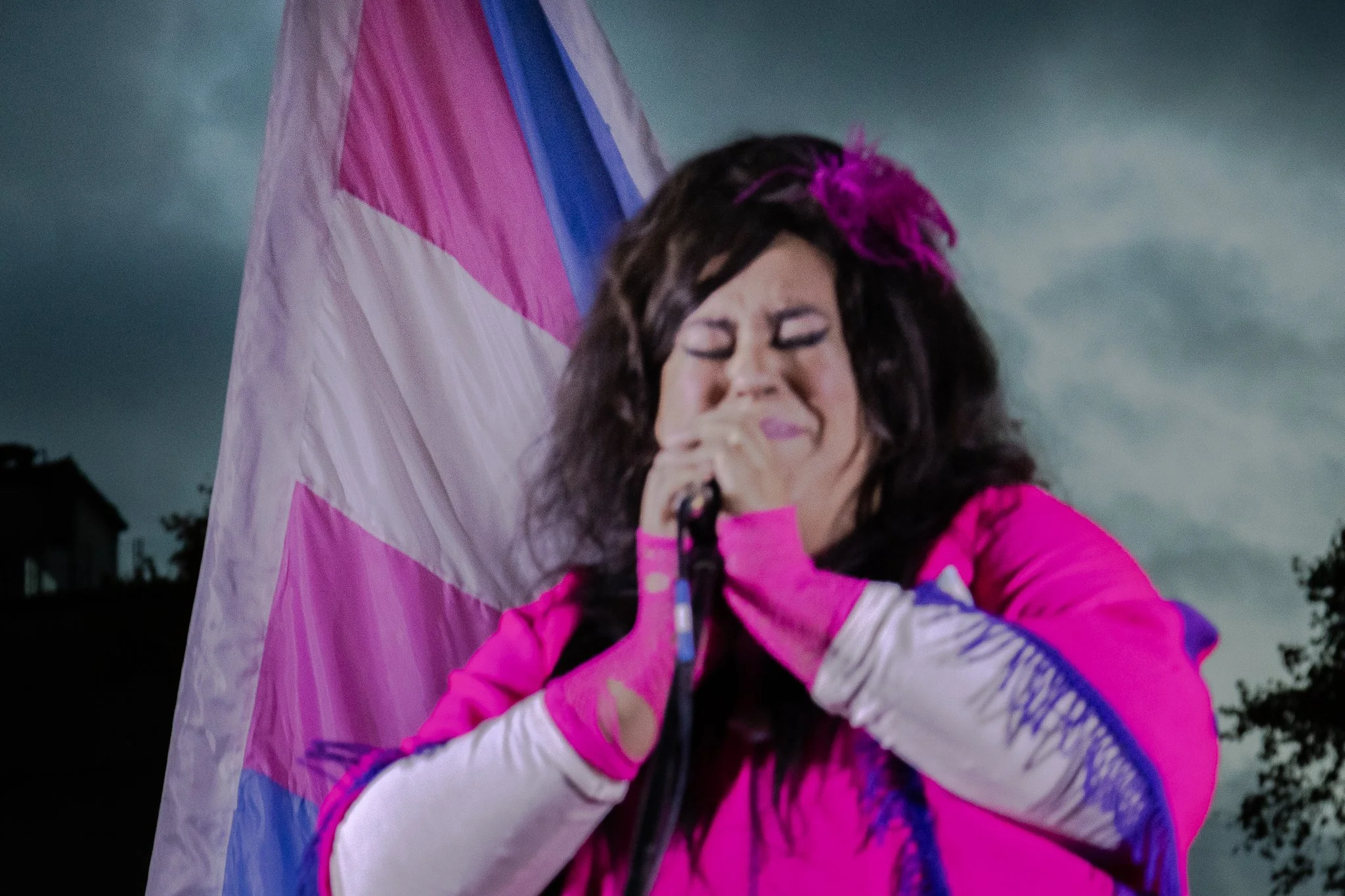 Mujer con cabello rizado, cantando con un micrófono y sosteniendo una bandera trans con los colores rosa, blanco y azul en un entorno al aire libre con cielo nublado.