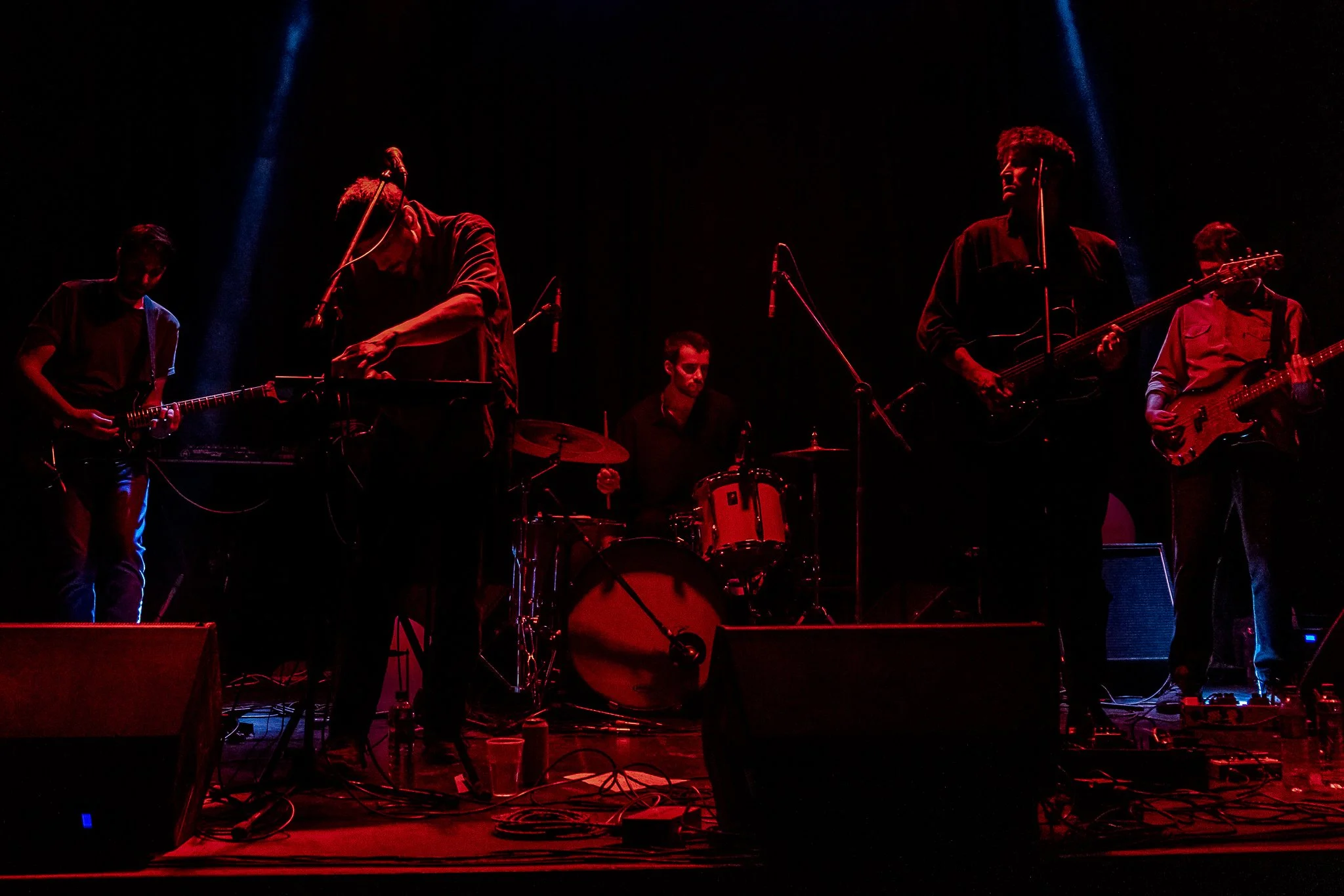 Un grupo de músicos tocando en un escenario oscuro, iluminados por luces rojas y azules, con instrumentos como guitarras, batería y micrófonos.