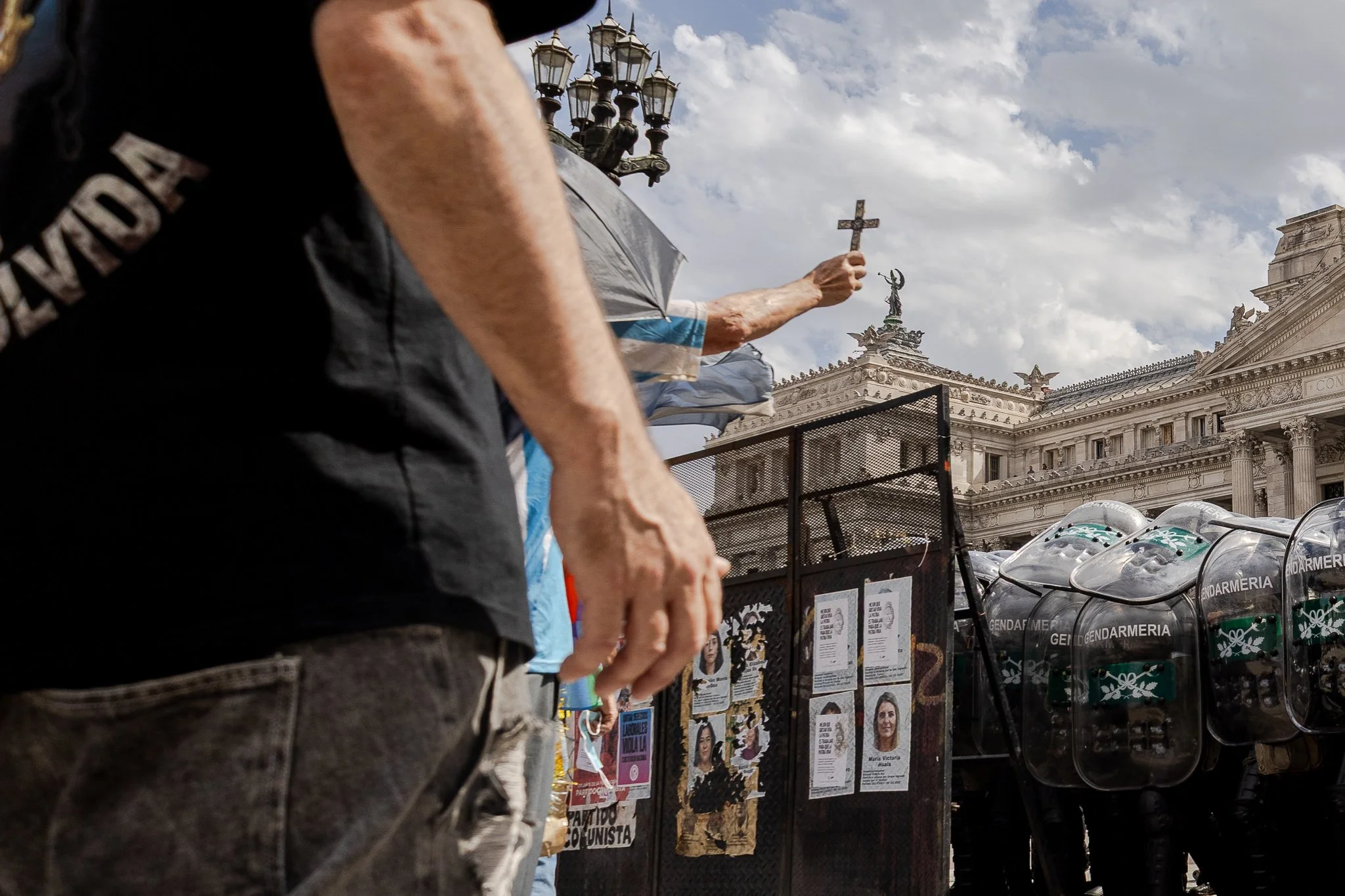 Personas participando en una manifestación frente a un edificio antiguo con arquitectura clásica y estatuas, sosteniendo cruces y banderas, con escudos antiprotesta y carteles en la calle.