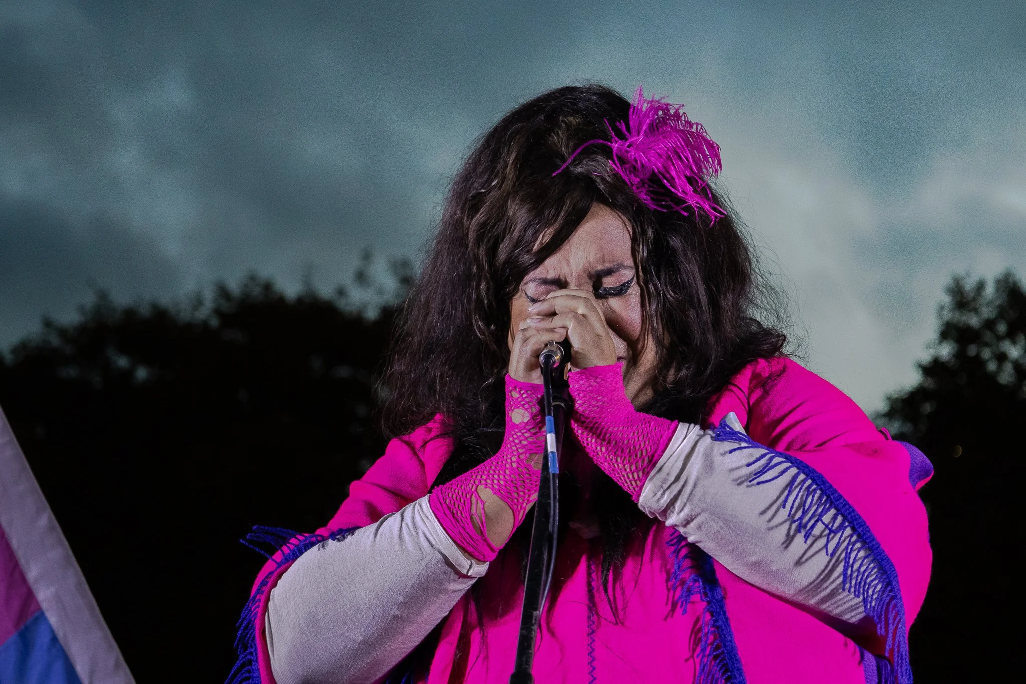 Mujer con micrófono, vestida con ropa rosa y con detalles azules, con pestañas largas y peinada con una cinta rosa y plumas. 