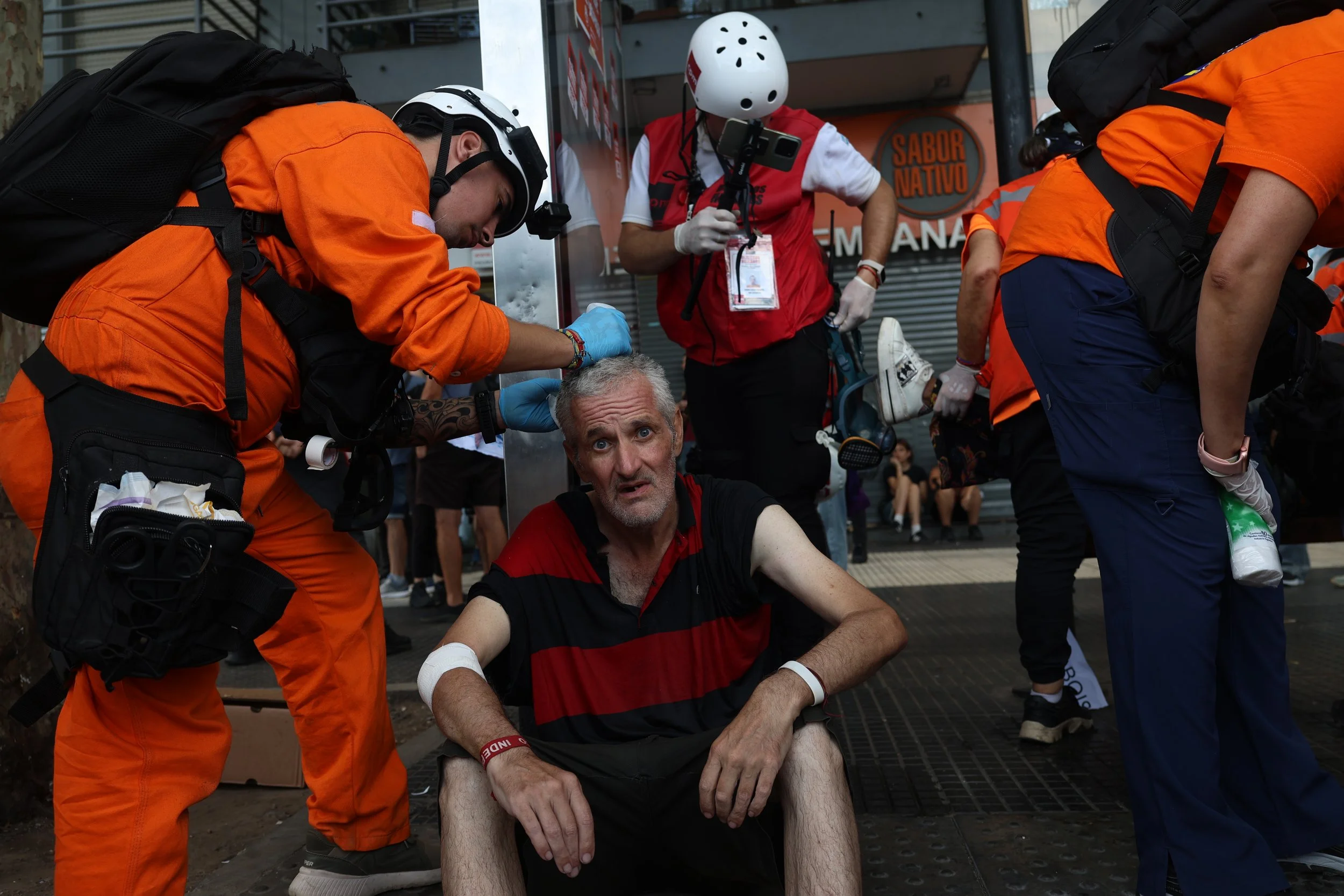 Botiquín de primeros auxilios en la calle con un hombre herido, rodeado de paramédicos con equipo de emergencia, algunos con cascos y uniformes naranjas y otros con uniformes de protección y mochilas.