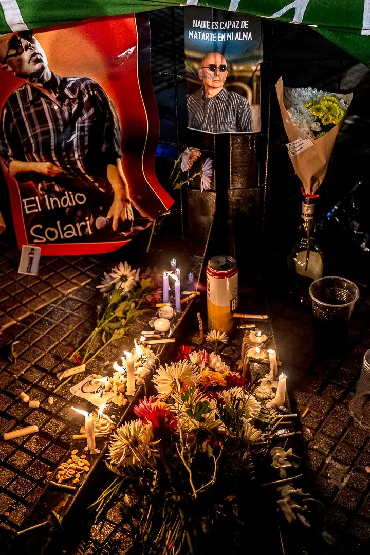 Un altar con flores, velas encendidas, y fotografías del Indio Solari. Incluye una caja de cerveza y una taza de plástico, con posters y flores como ofrendas.