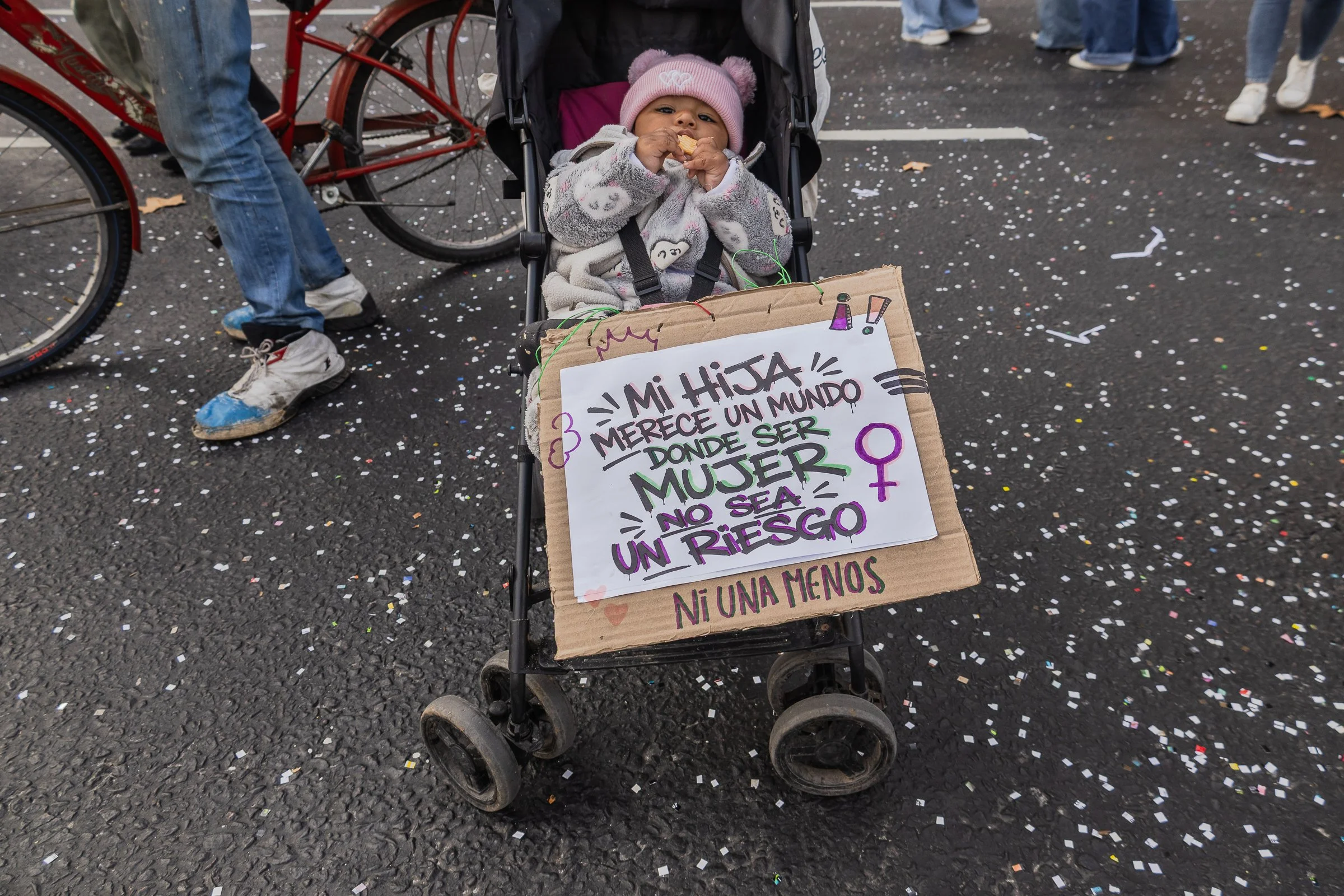 Bebé en un cochecito, con gorro rosado, comiendo galleta, en una marcha con confeti en el suelo, el cochecito tiene un cartel decorativo que dice 'Mi hija merece un mundo donde ser mujer no sea un riesgo. Ni una menos'.
