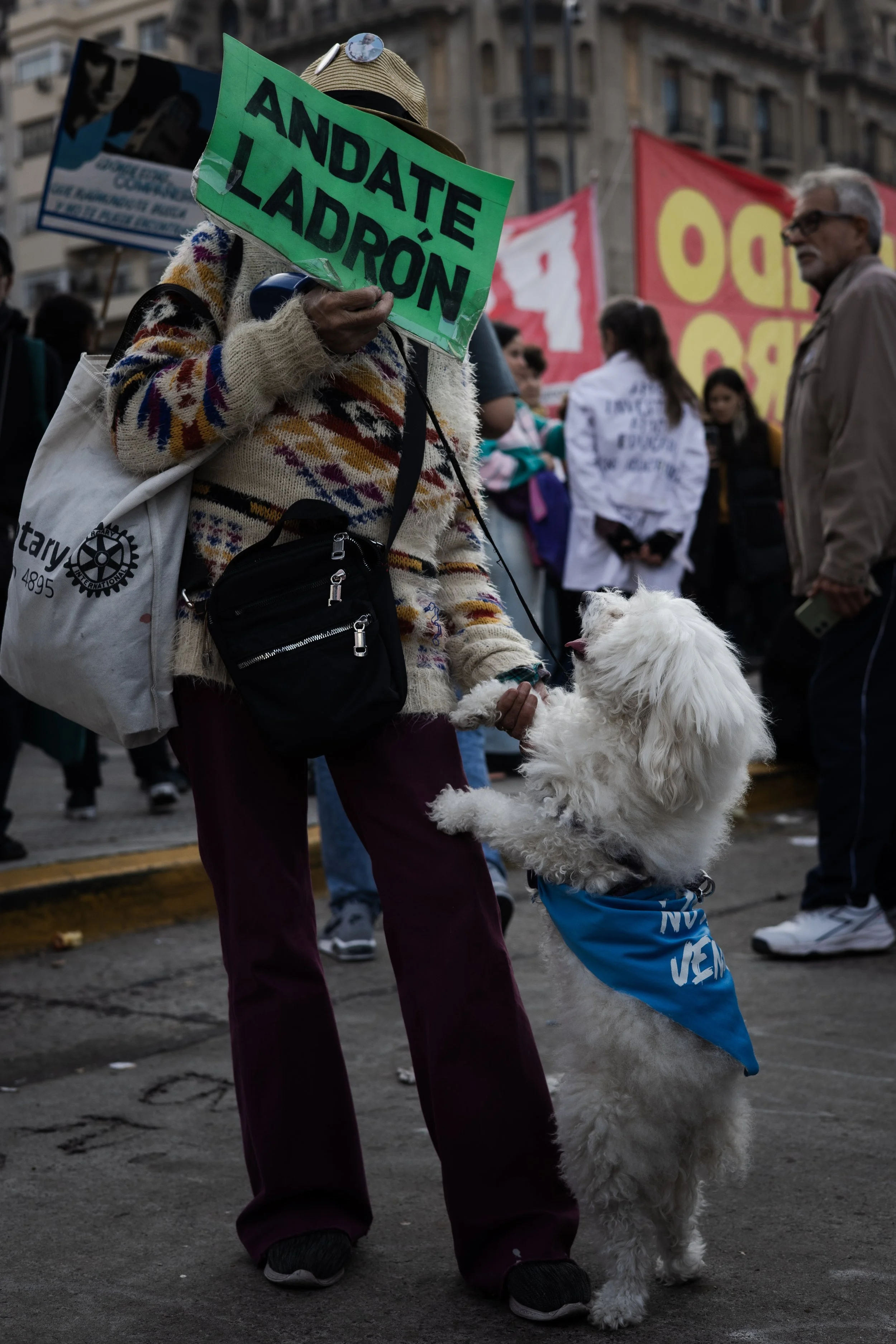 Persona sosteniendo una señal que dice 'Andate Ladron' en una protesta, acompañada de un perro con bandana azul que se jala de la mano de la persona.