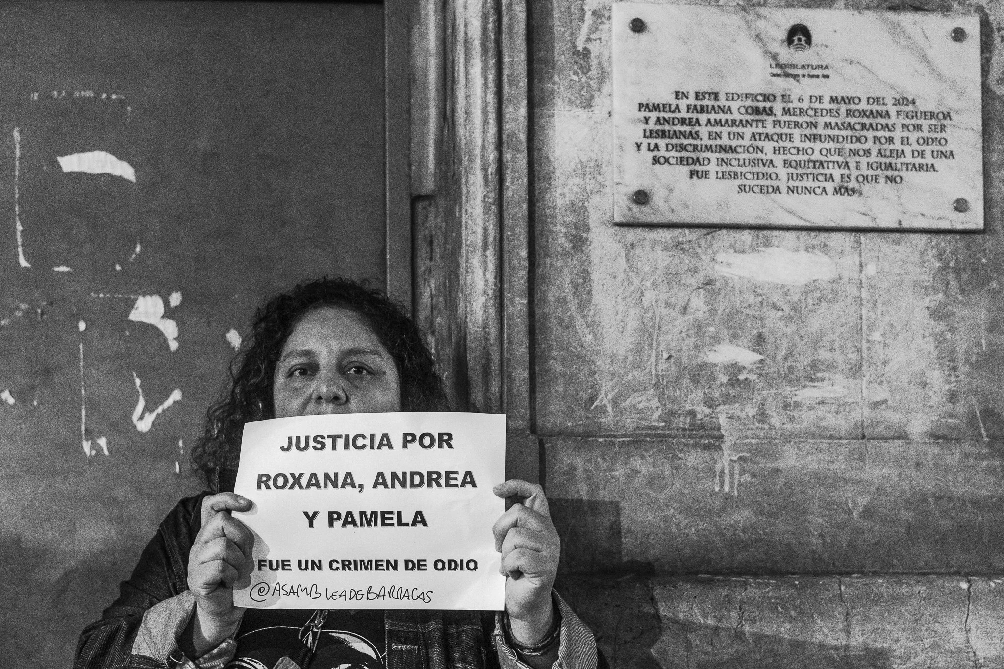 Mujer sosteniendo un cartel que dice "JUSTICIA POR ROXANA, ANDREA Y PAMELA" en un entorno urbano con paredes desgastadas.