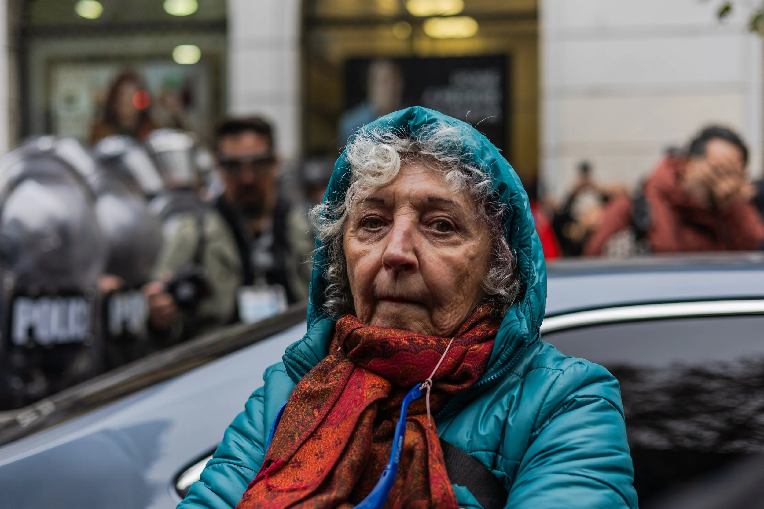 Mujer mayor con chaqueta azul y bufanda naranja en la calle, con manifestantes y policías antimotines en el fondo.