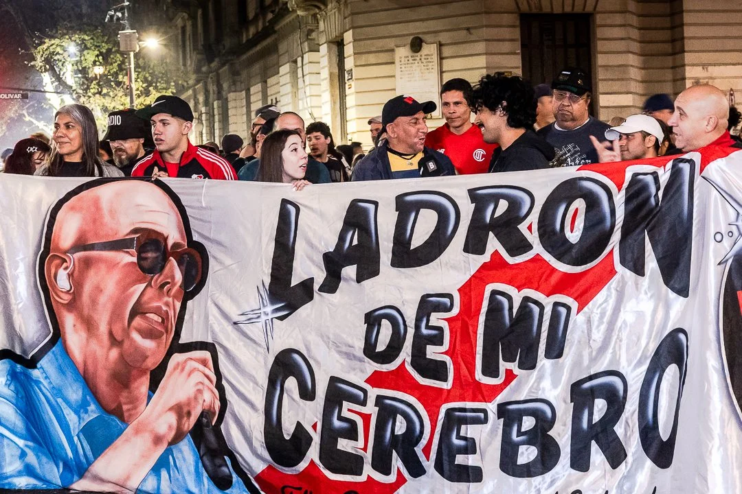 Grupo de personas sosteniendo una pancarta con una caricatura del Indio Solari y el texto "LADRON DE MI CEREBRO" en un ambiente nocturno en la calle.