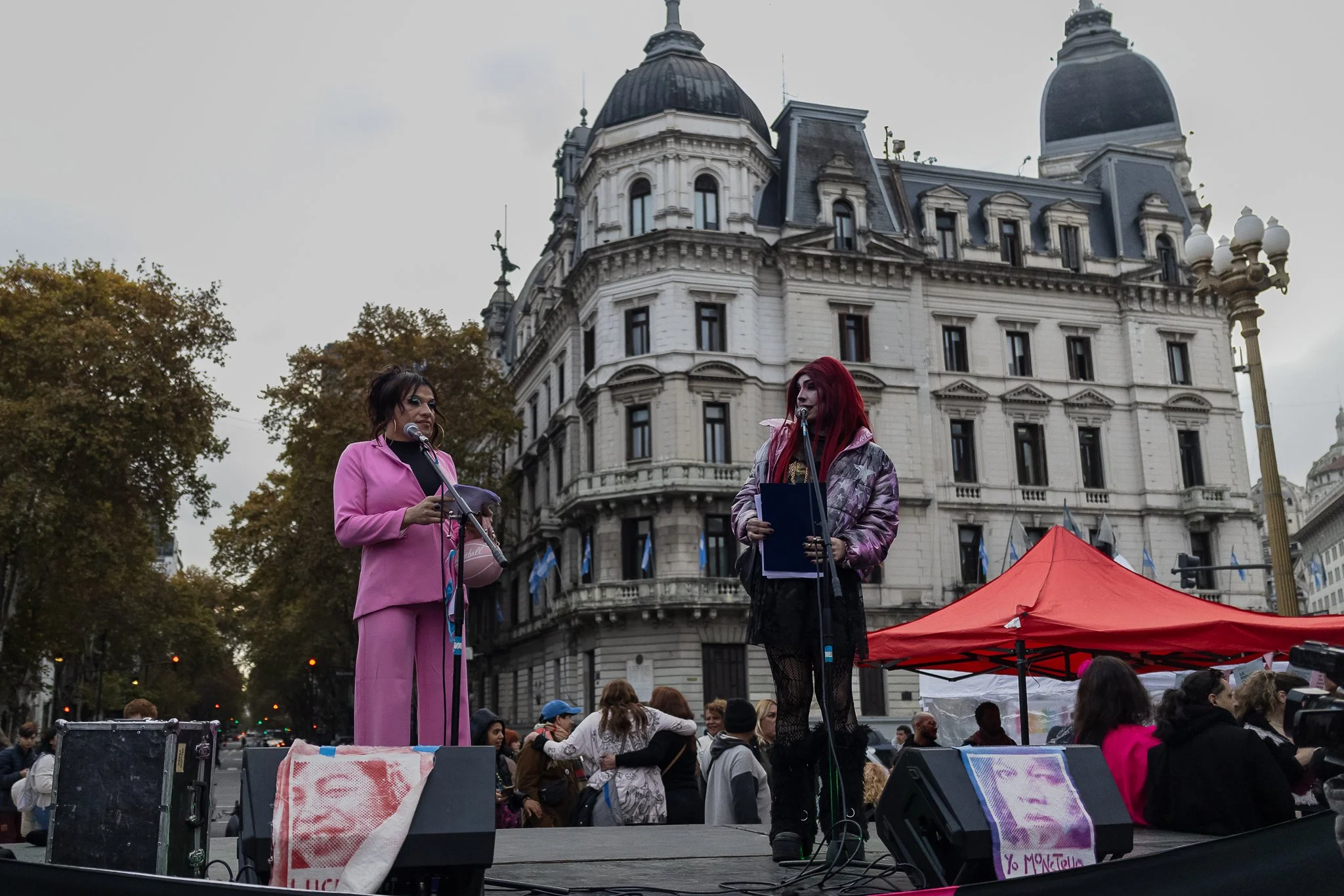 Dos mujeres en un escenario durante una protesta en una ciudad, con un edificio histórico de fondo y un grupo de personas en el público.