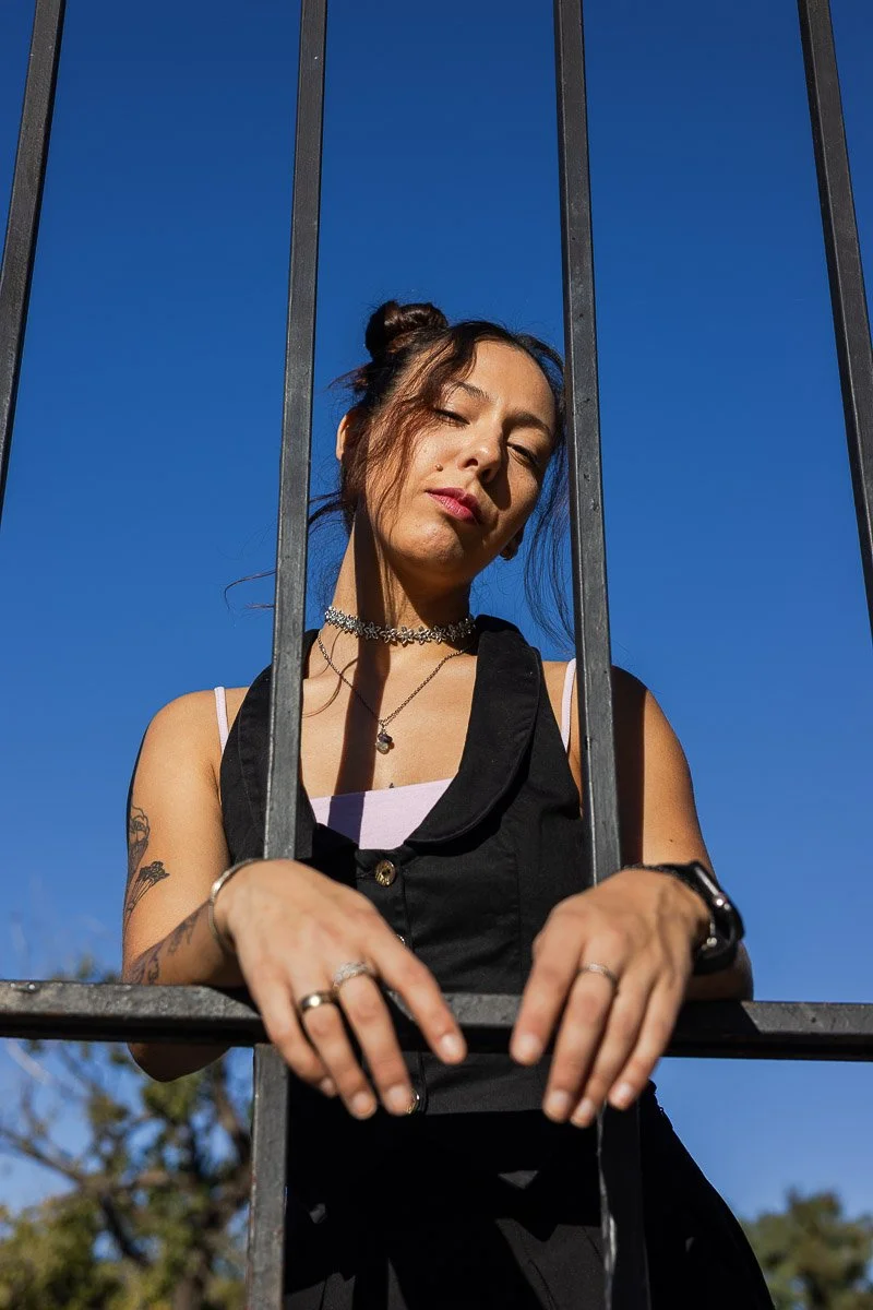 Juventud femenina con maquillaje, cabello en doble coleta, sosteniendo una reja metálica en un día soleado con cielo azul.