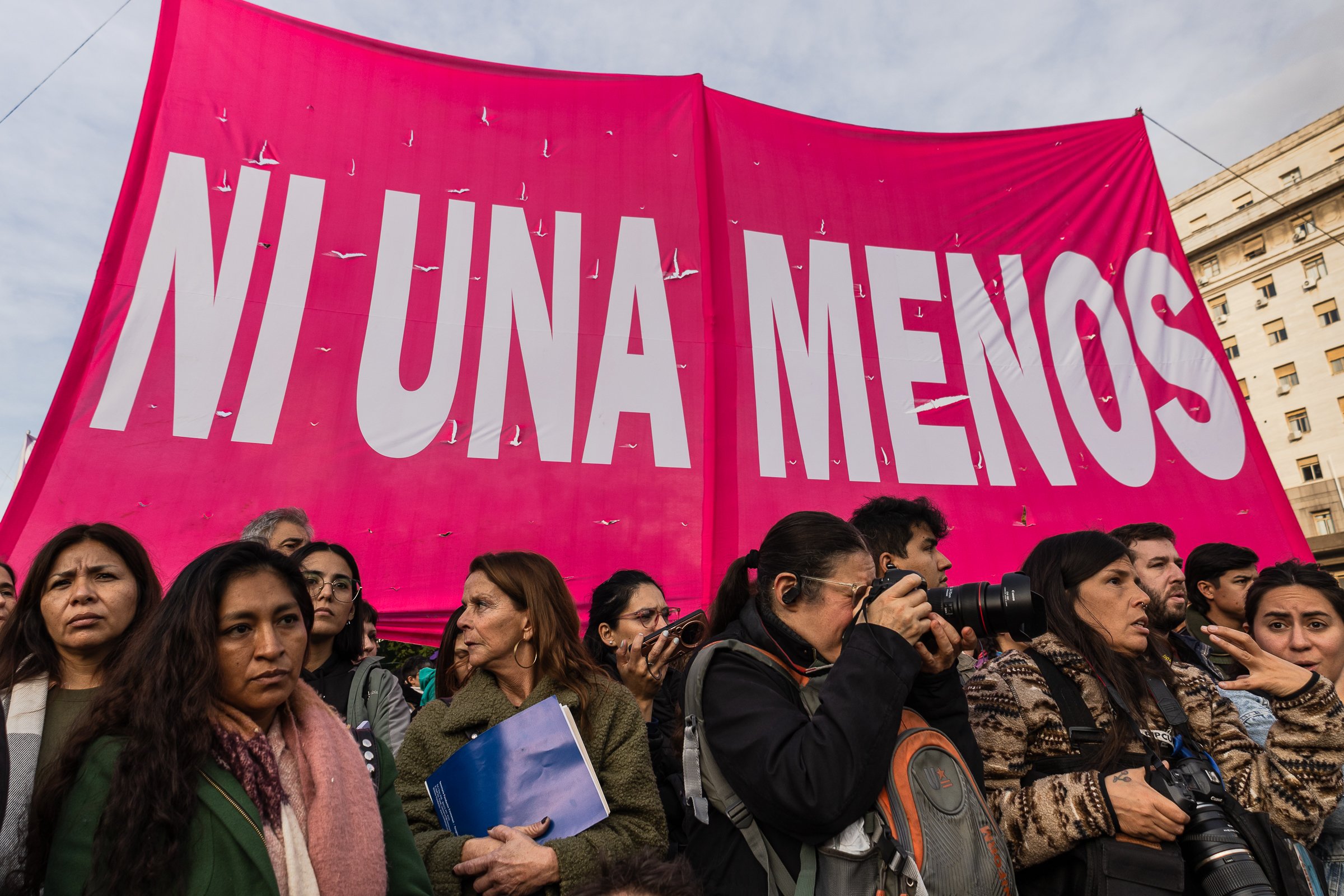 Grupo de personas en una manifestación con cartel rosa que dice 'Ni una menos' en letras blancas, en un entorno urbano durante el día.