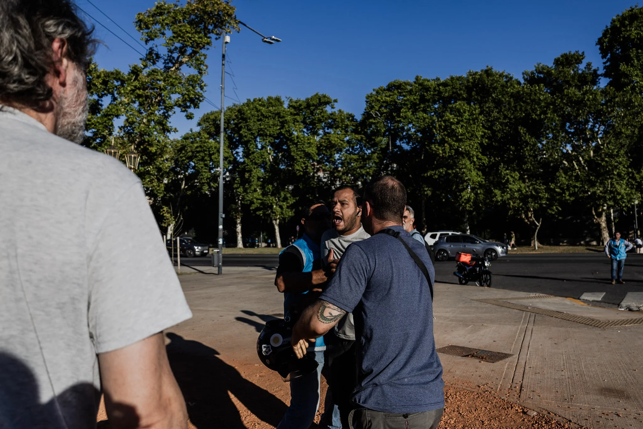 Varias personas involucradas en un altercado en la vía pública, con fondo de árboles y autos en una ciudad.