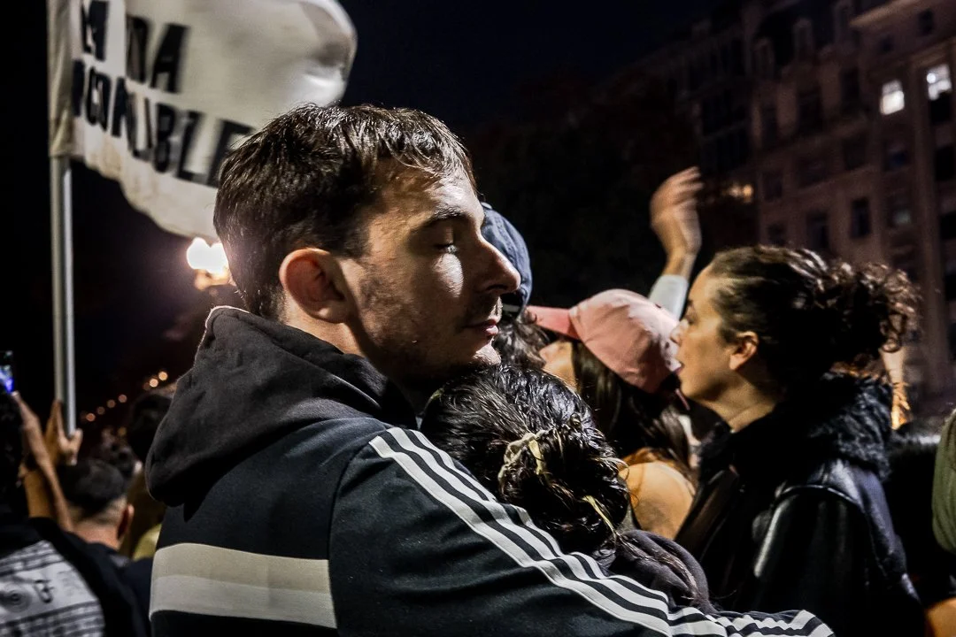 Joven con los ojos cerrados abrazando a otra chica, en medio de una multitud al atardecer en la calle.