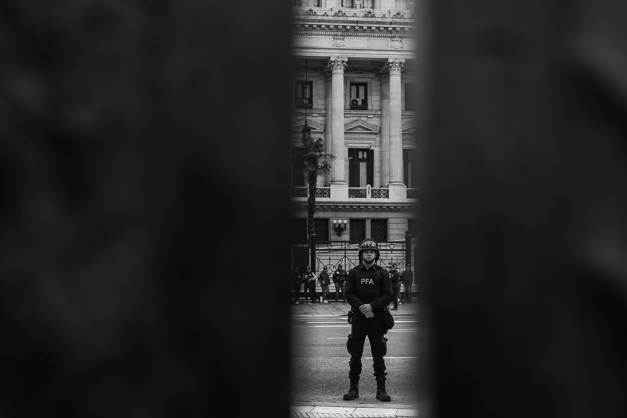 Un policía con uniforme y casco que tiene las letras 'PFA' en el pecho, de pie en medio de la calle en una zona urbana, con varios oficiales de policía en formación en el fondo y un edificio de estilo clásico con columnas, en la ciudad.