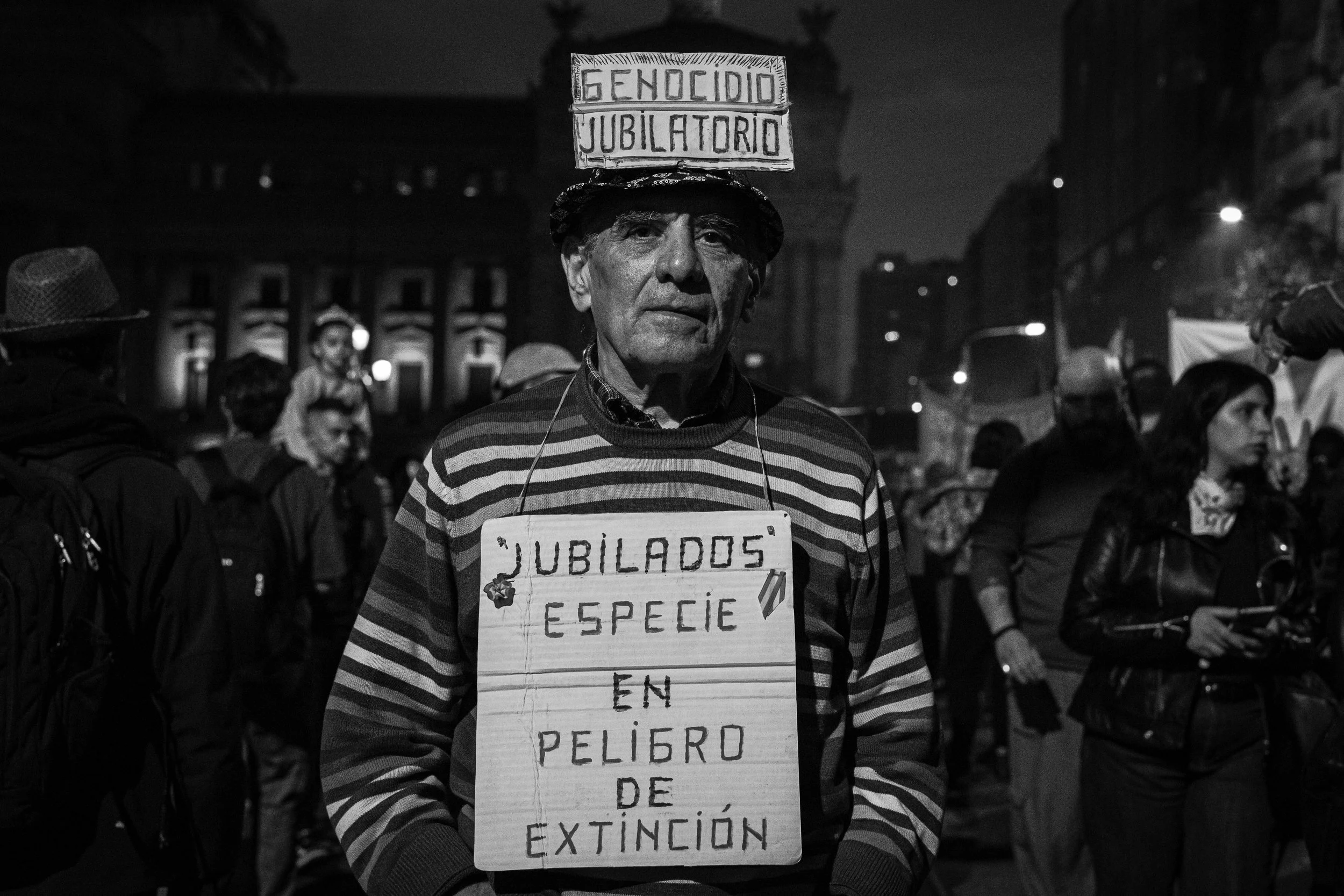 Hombre con camiseta a rayas y sombrero porta cartel que dice 'Jubilados en peligro de extinción', en una manifestación nocturna rodeado de otras personas.