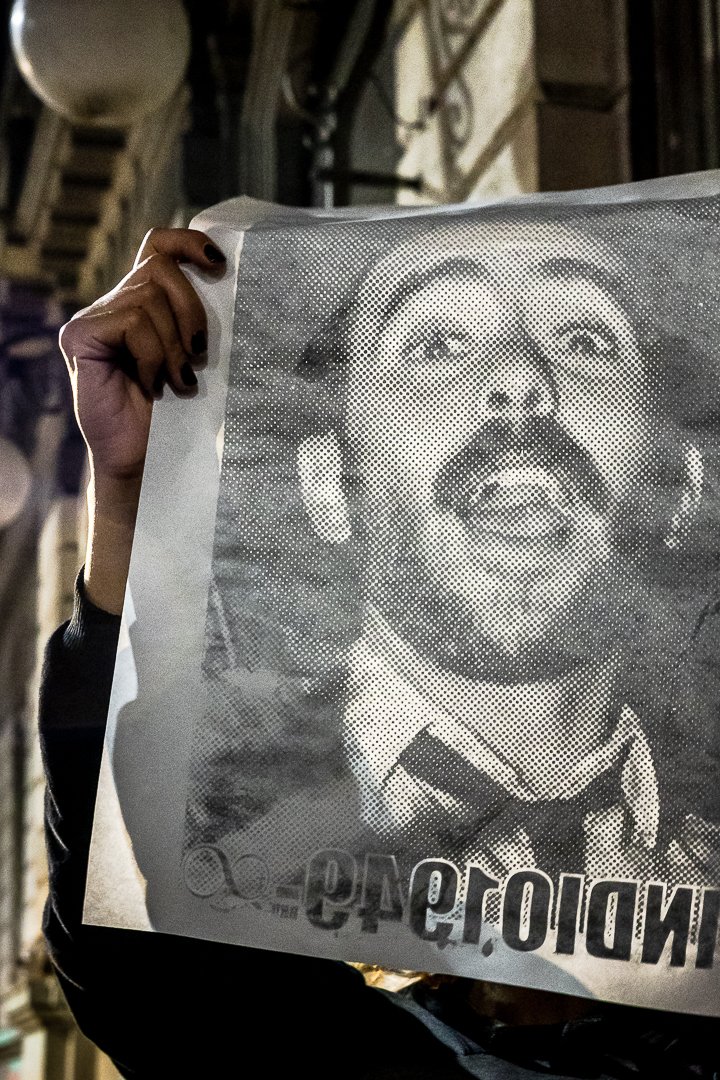 Persona sosteniendo un cartel con la imagen en blanco y negro de un hombre con barba, bigote y cabelo rizado, con expresión animada y boca abierta. El texto en el cartel está parcialmente visible.