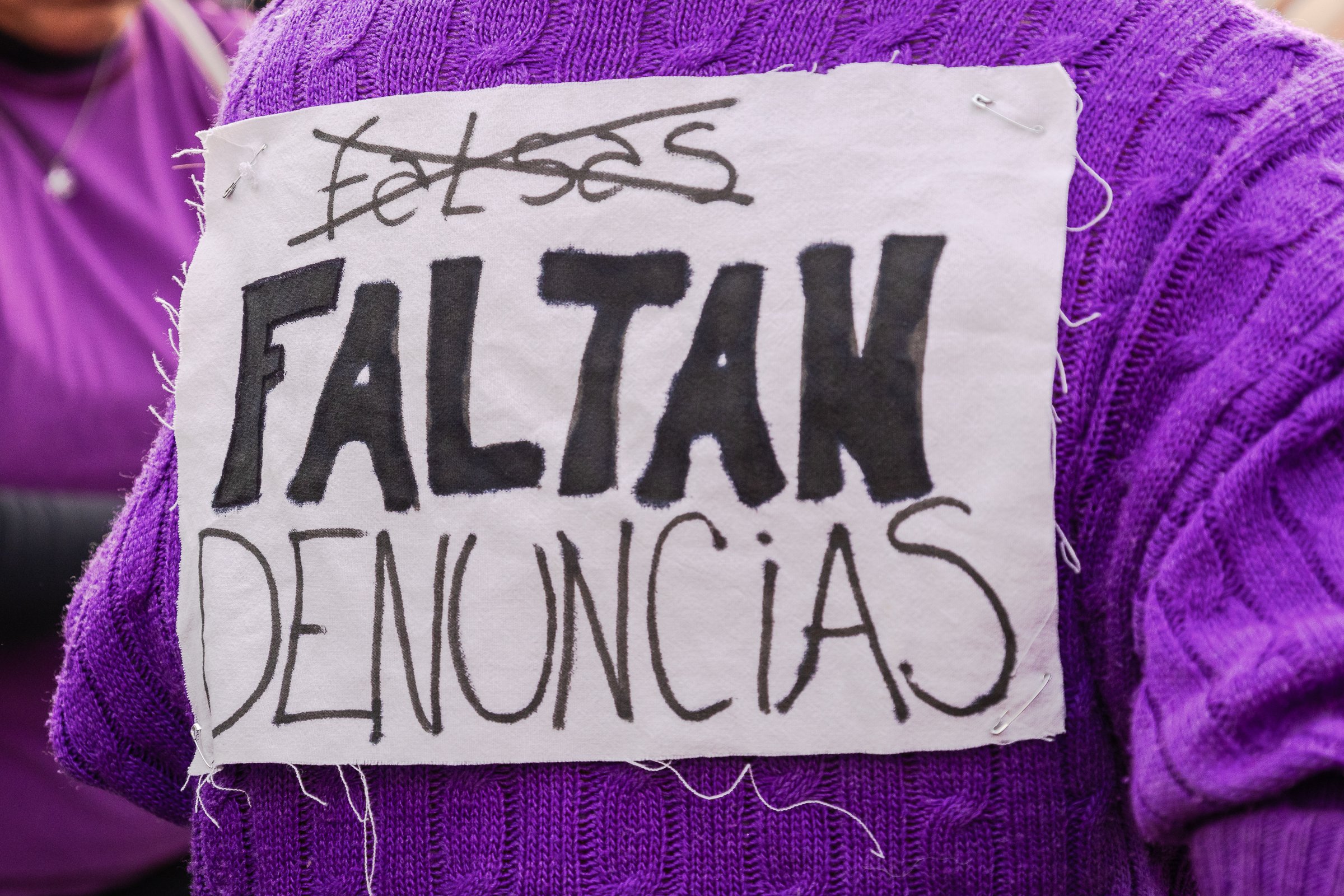 Persona con un suéter morado lleva un cartel con palabras en español que dice 'FALTAN DENUNCIAS'.