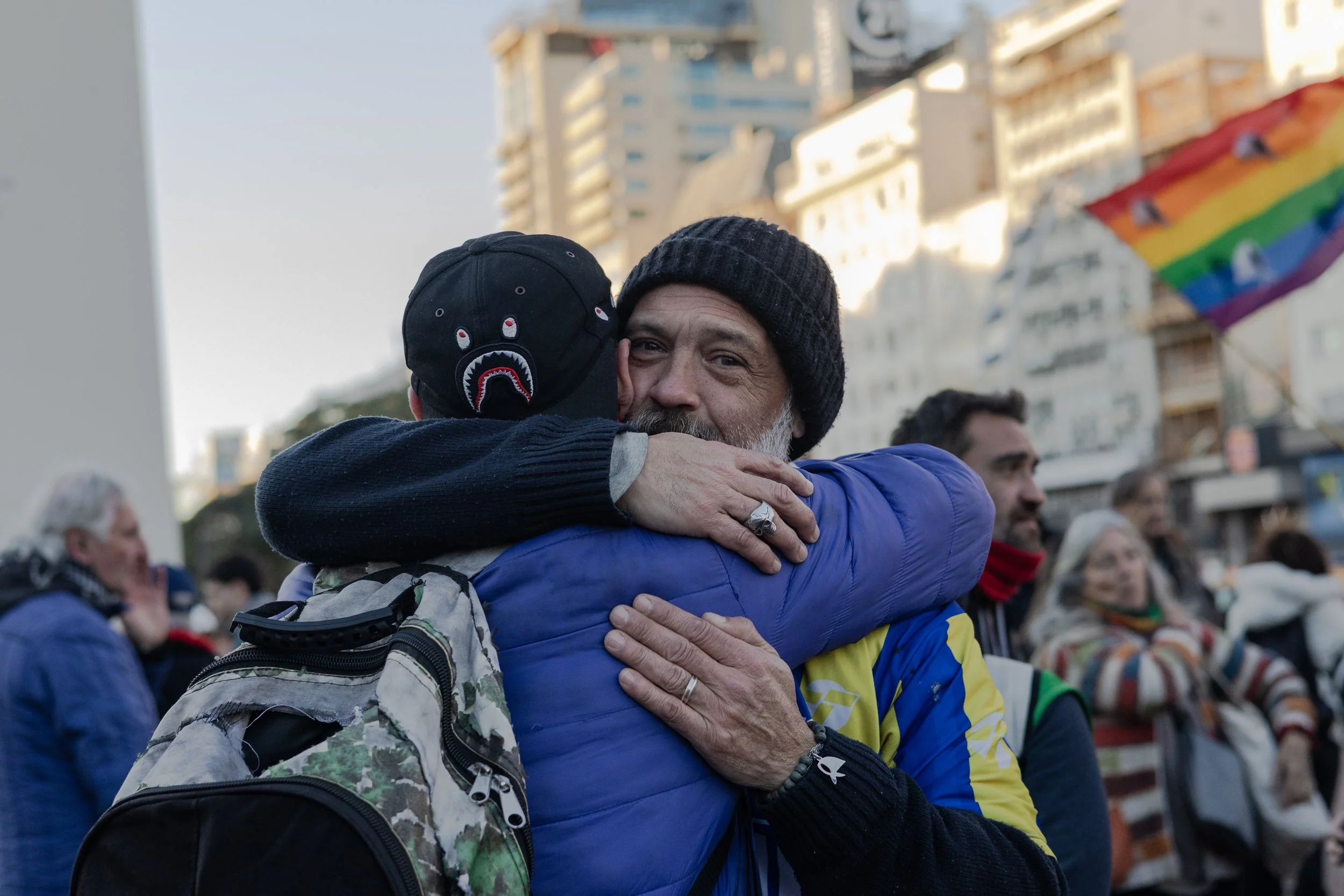Dos personas abrazándose en una reunión al aire libre, con varias otras personas y un arcoíris transgénero ondeando en fondo urbano.