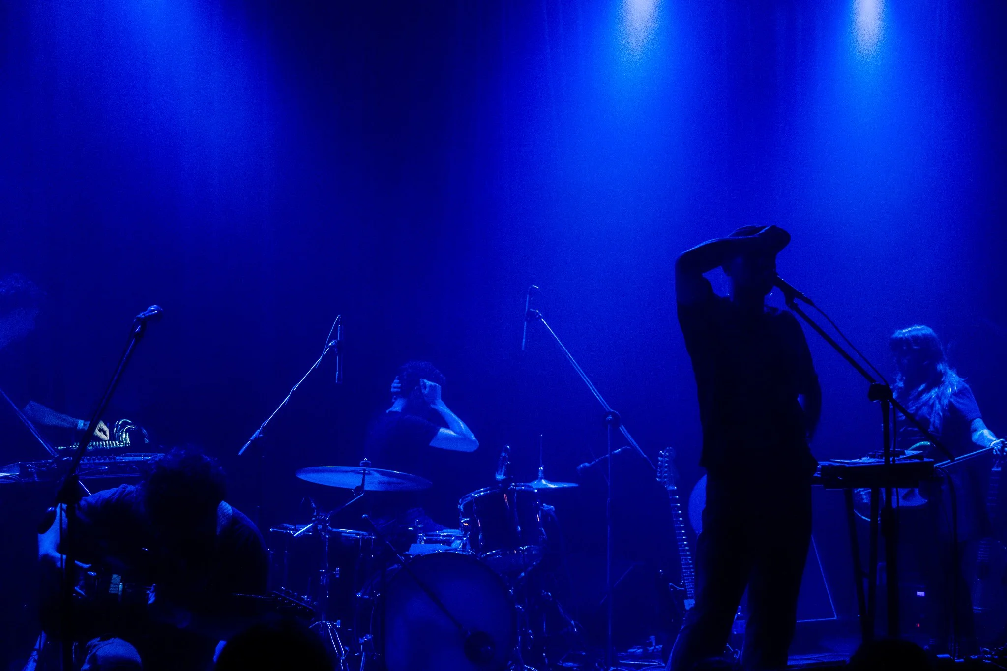 Músicos en un concierto, con iluminación azul, algunos tocando instrumentos y uno ajustando su micrófono.