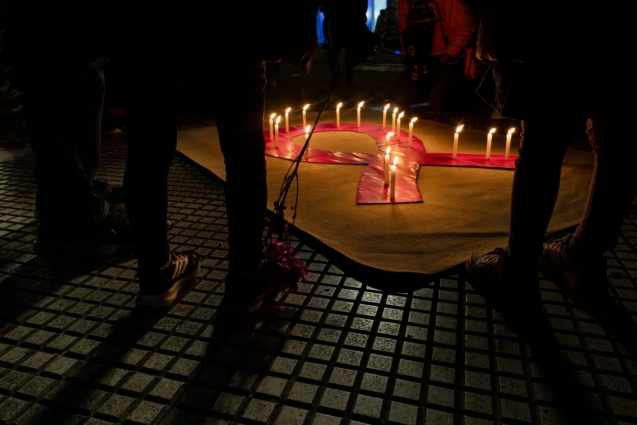 Personas rodeando unarojilla con velas encendidas formando un símbolo de rosa, participando en una ceremonia o acto conmemorativo en la noche.