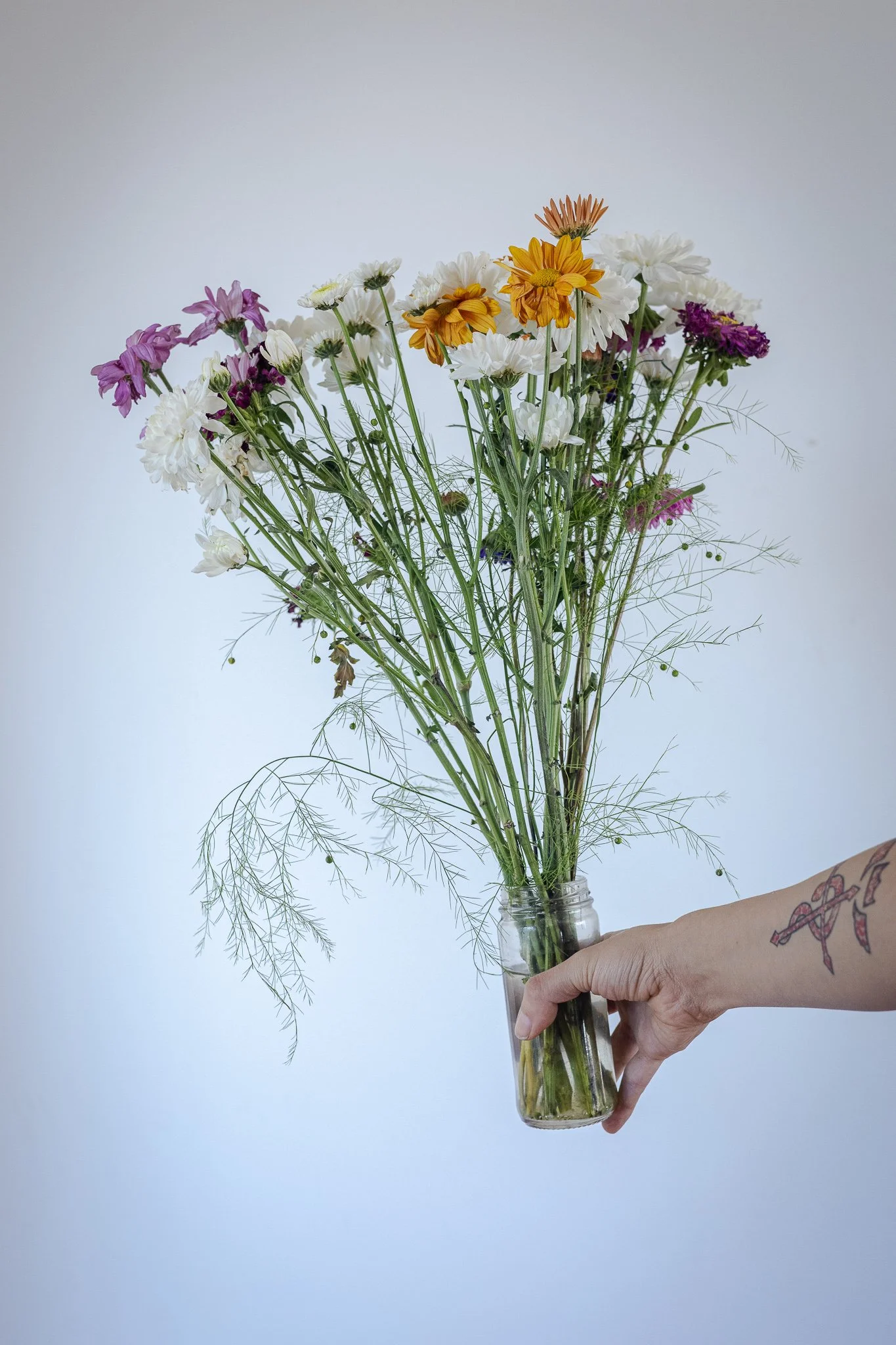 Ramo de flores variadas en un vaso de vidrio, sostenido por una mano con tatuaje.
