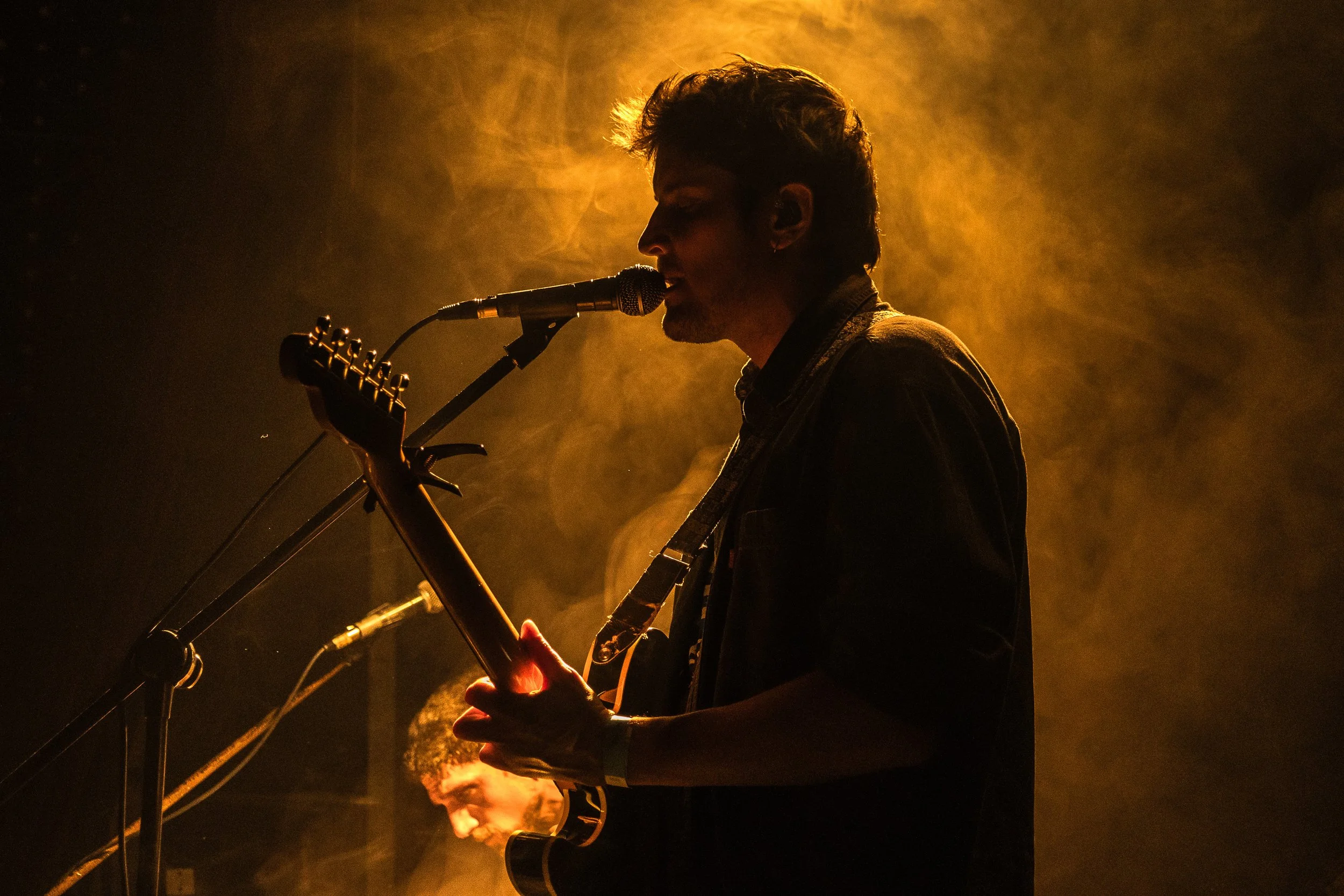 Un hombre tocando la guitarra y cantando en un escenario con iluminación cálida y humo.
