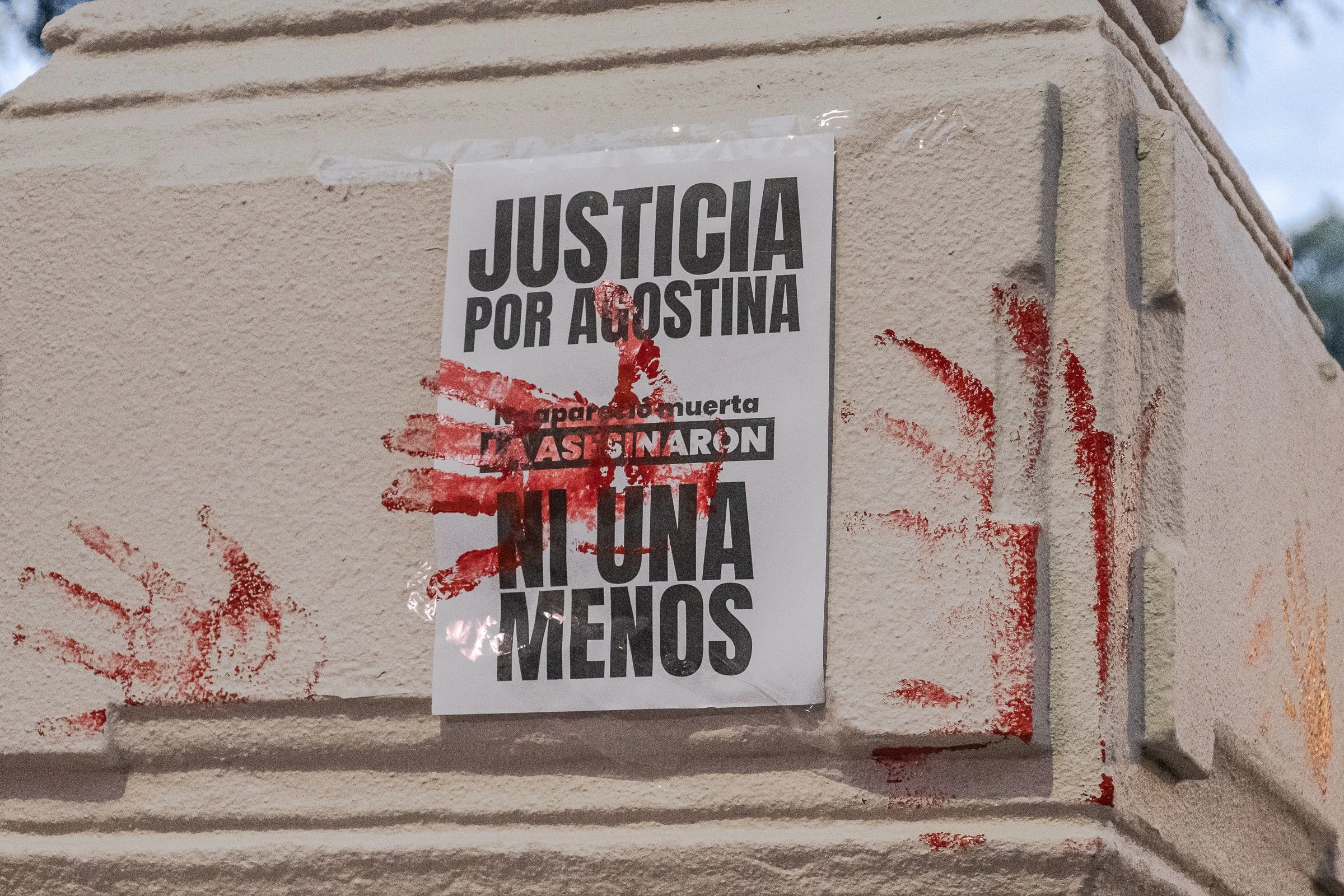 Papel con la frase 'Justicia por Agostina' pegado en una estructura de piedra, con manchas de pintura roja alrededor.