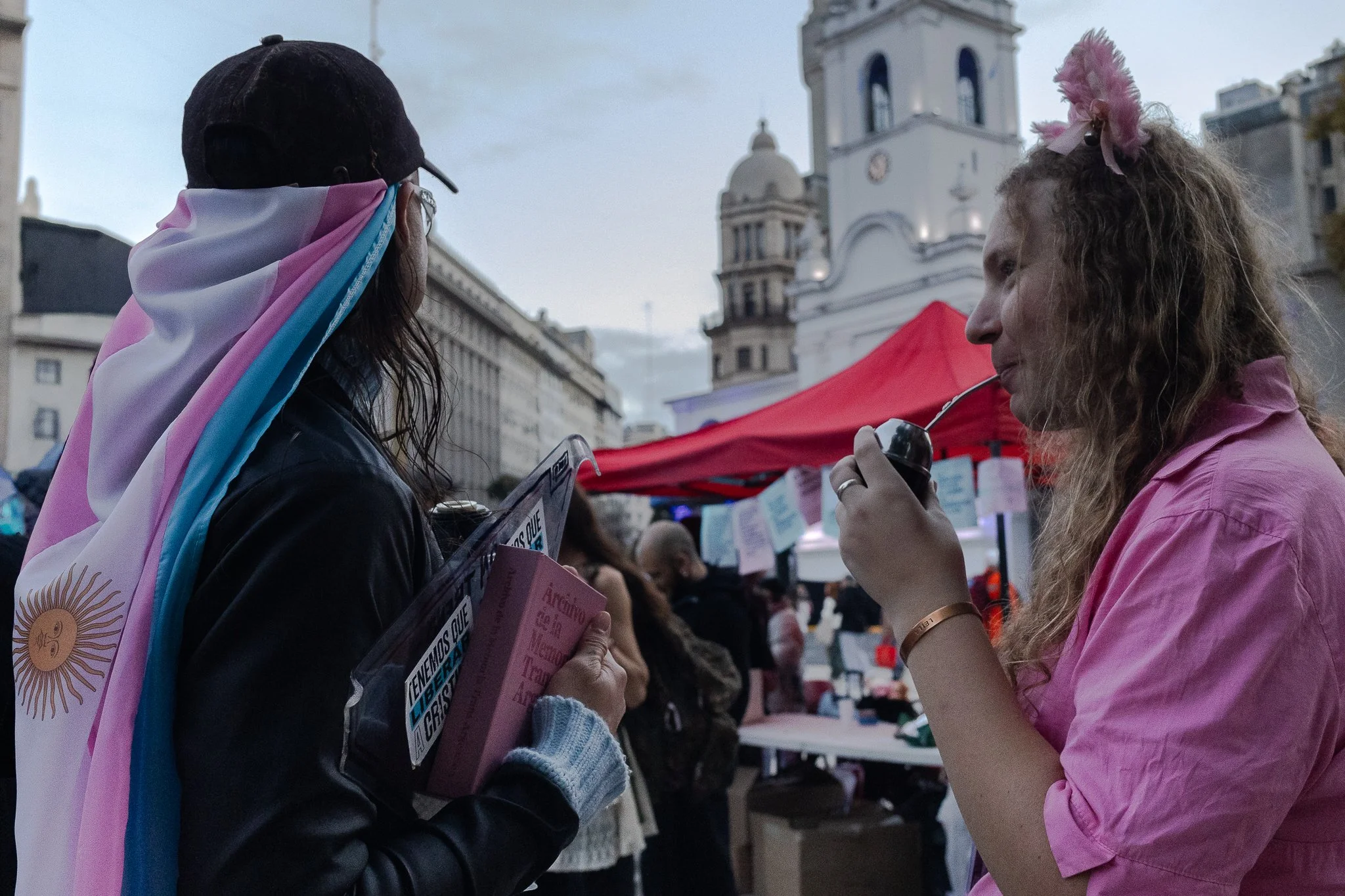 Dos mujeres conversando en una plaza en un evento, una lleva una bandera de Argentina y trans, la otra sostiene un mate. Al fondo, edificios y una estructura de iglesia con reloj.