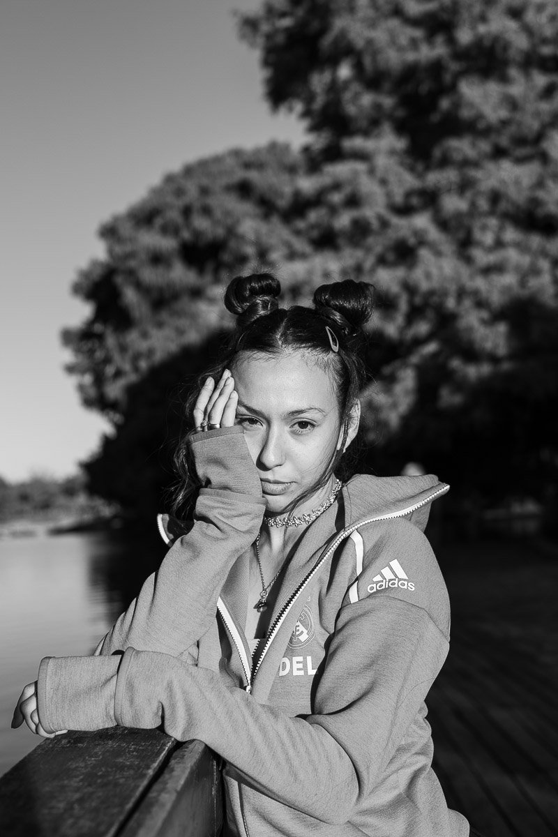 Joven mujer con cabello oscuro en dos moños, usando chaqueta deportiva con logotipo de Adidas, posando en un entorno natural junto a un río con árboles en el fondo.
